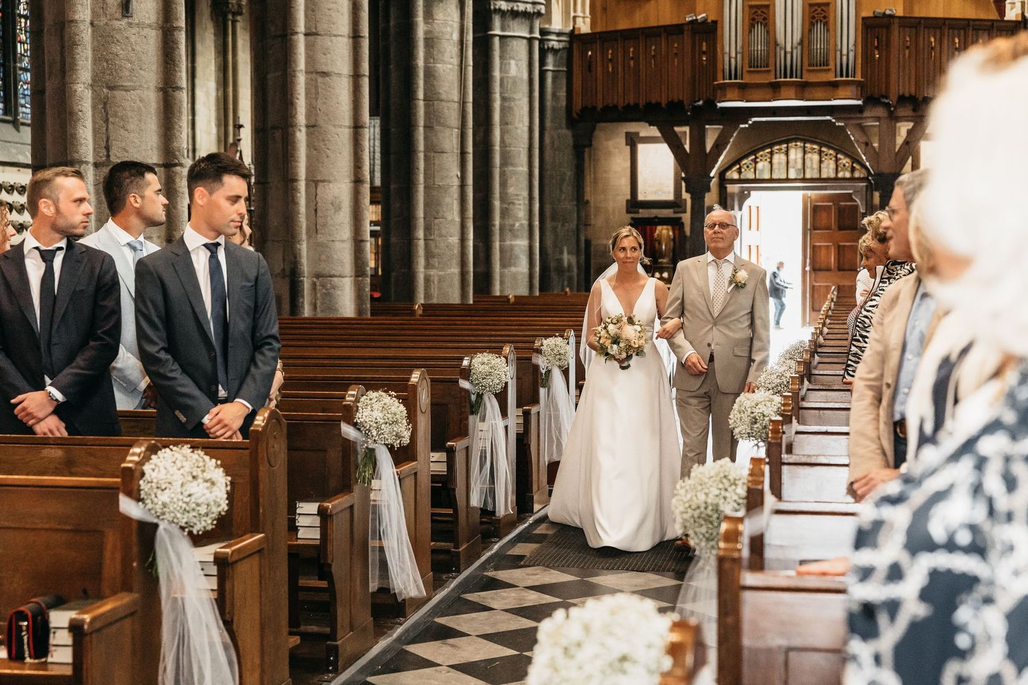 Bride walks down cathedral aisle with father past white floral pew decorations toward waiting groomsmen.