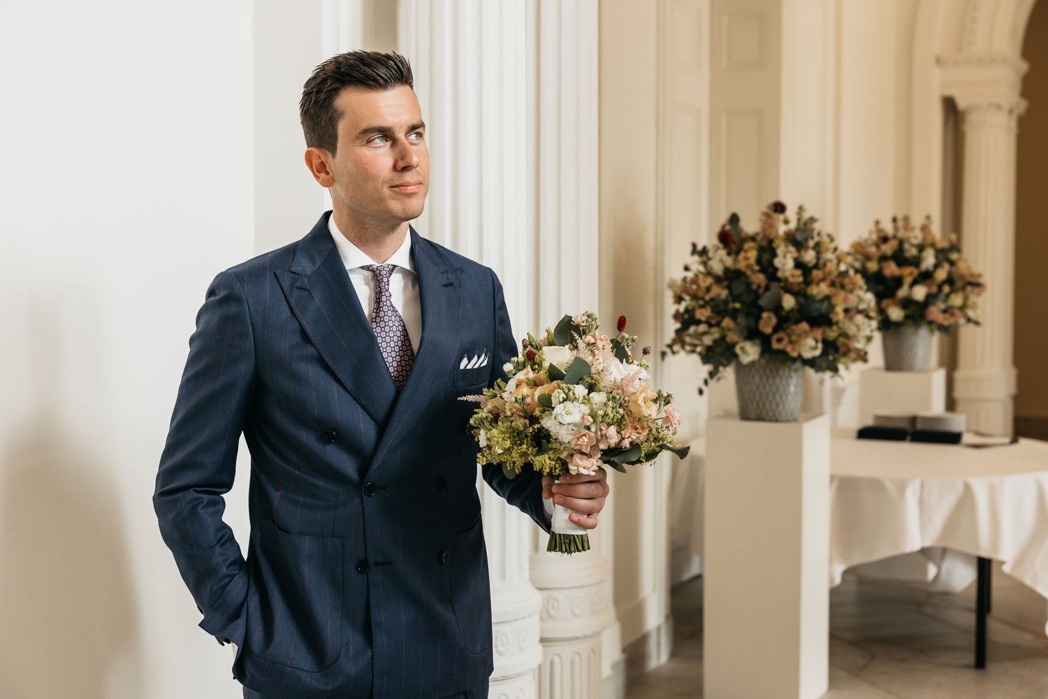 Groom in navy suit holding bridal bouquet of roses awaiting bride at elegant wedding venue.