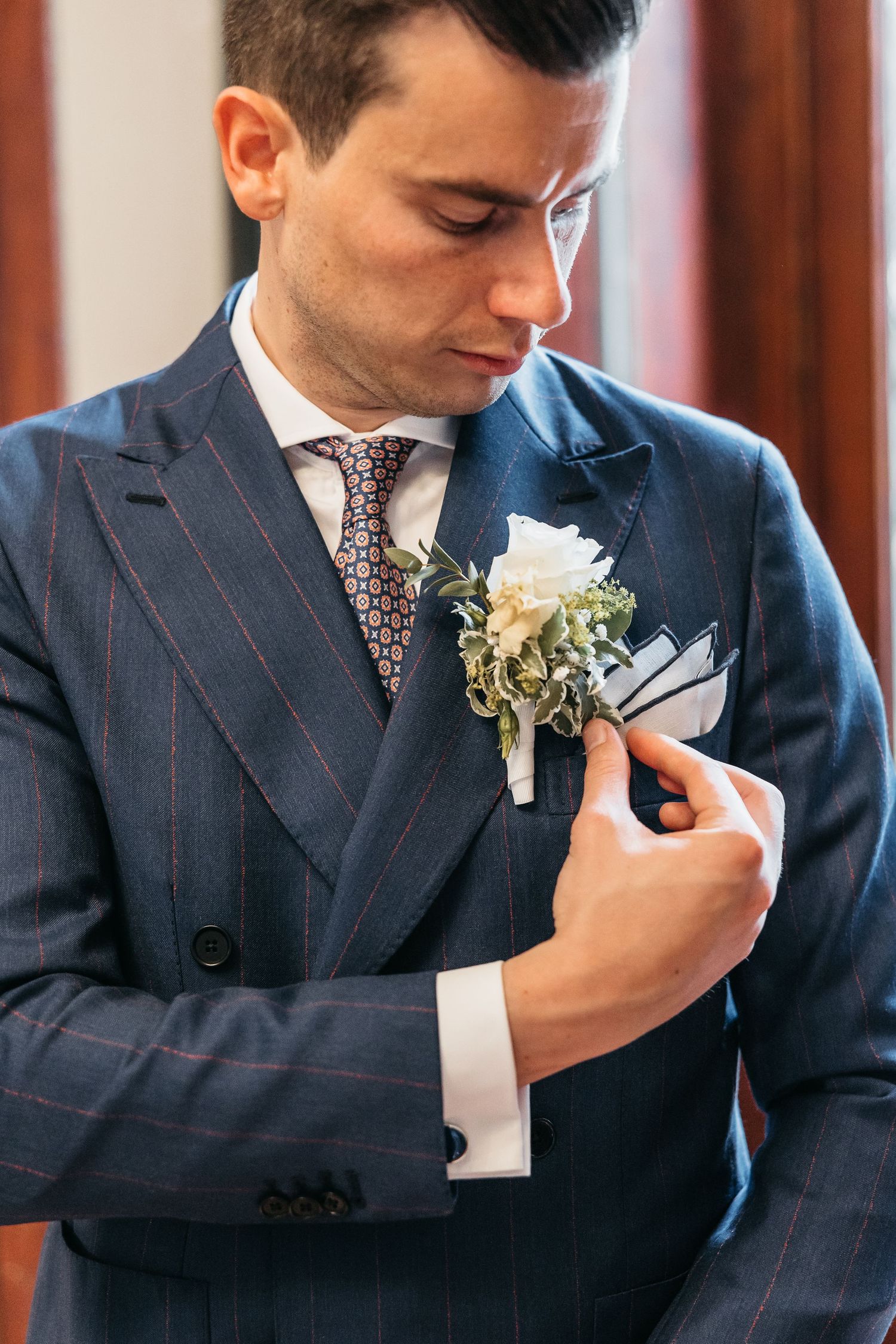 Dapper groom in navy pinstripe suit adjusting white floral boutonniere with patterned tie and pocket square.