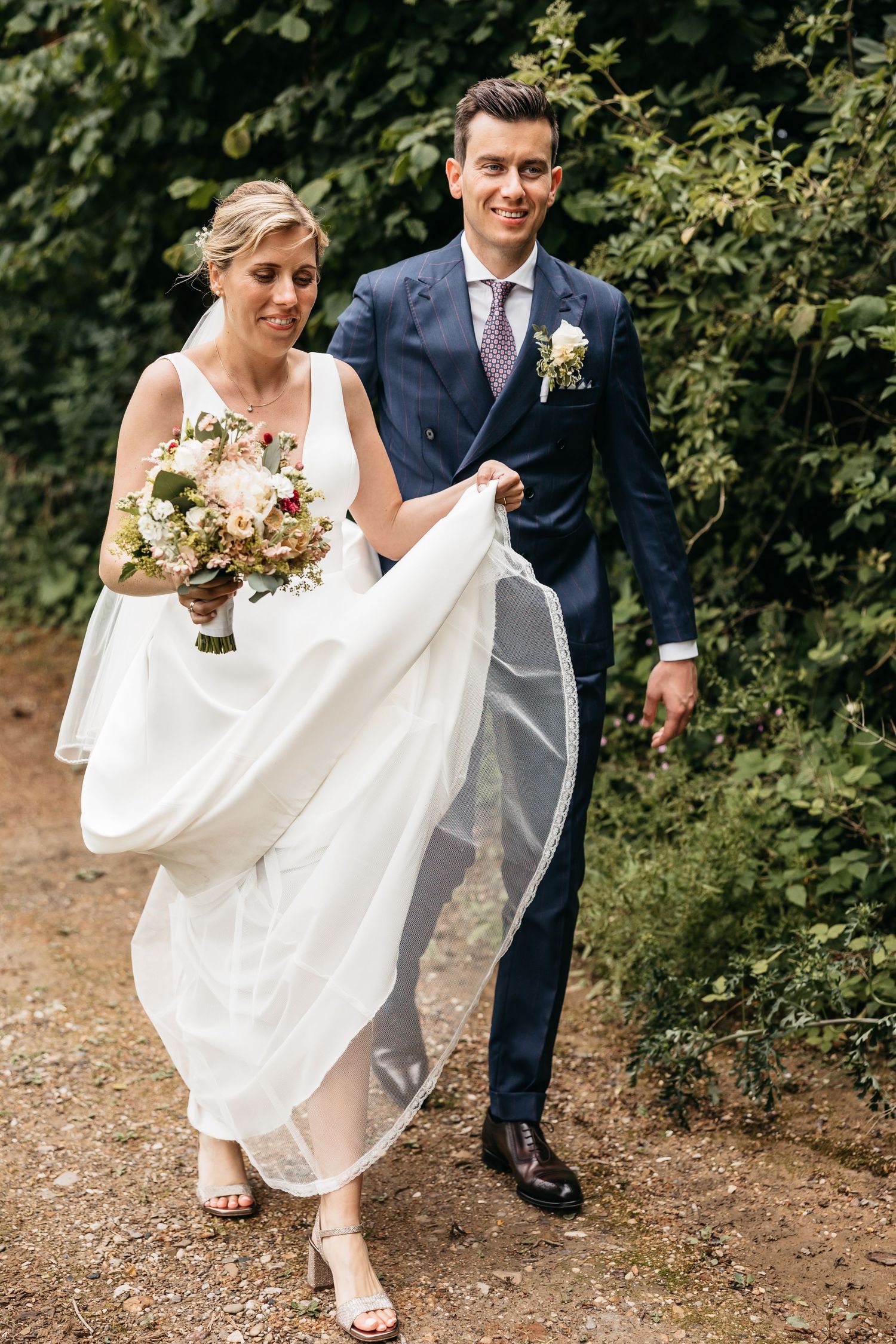 Smiling bride and groom walking hand in hand along garden path, bride holding bouquet.