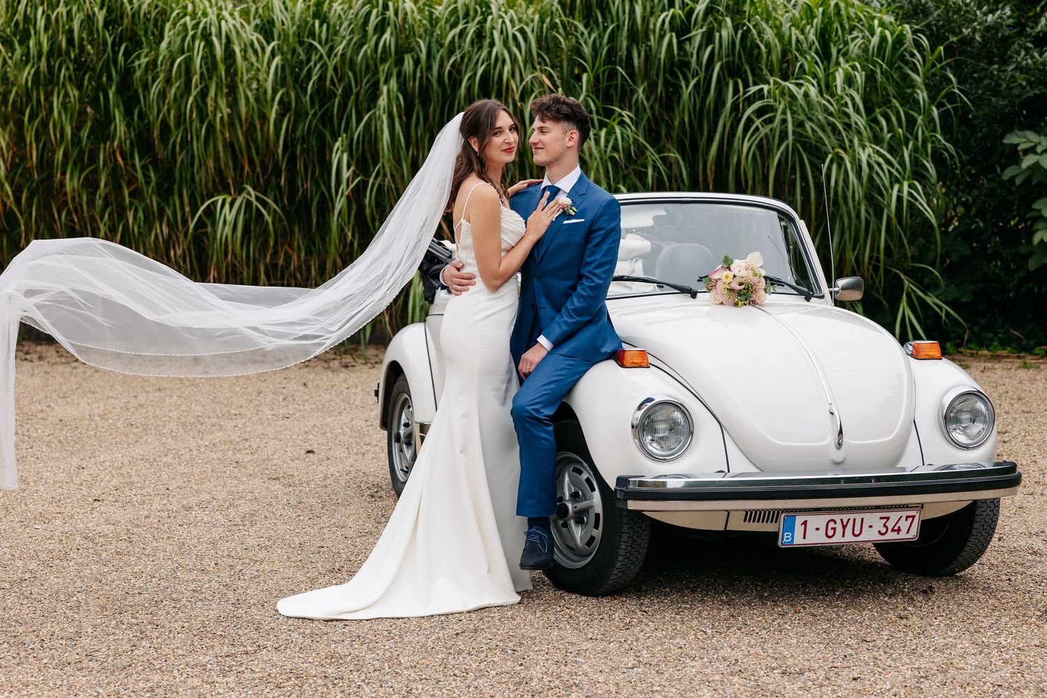 Bride and groom embrace beside a classic white Volkswagen Beetle convertible with veil flowing in the breeze.