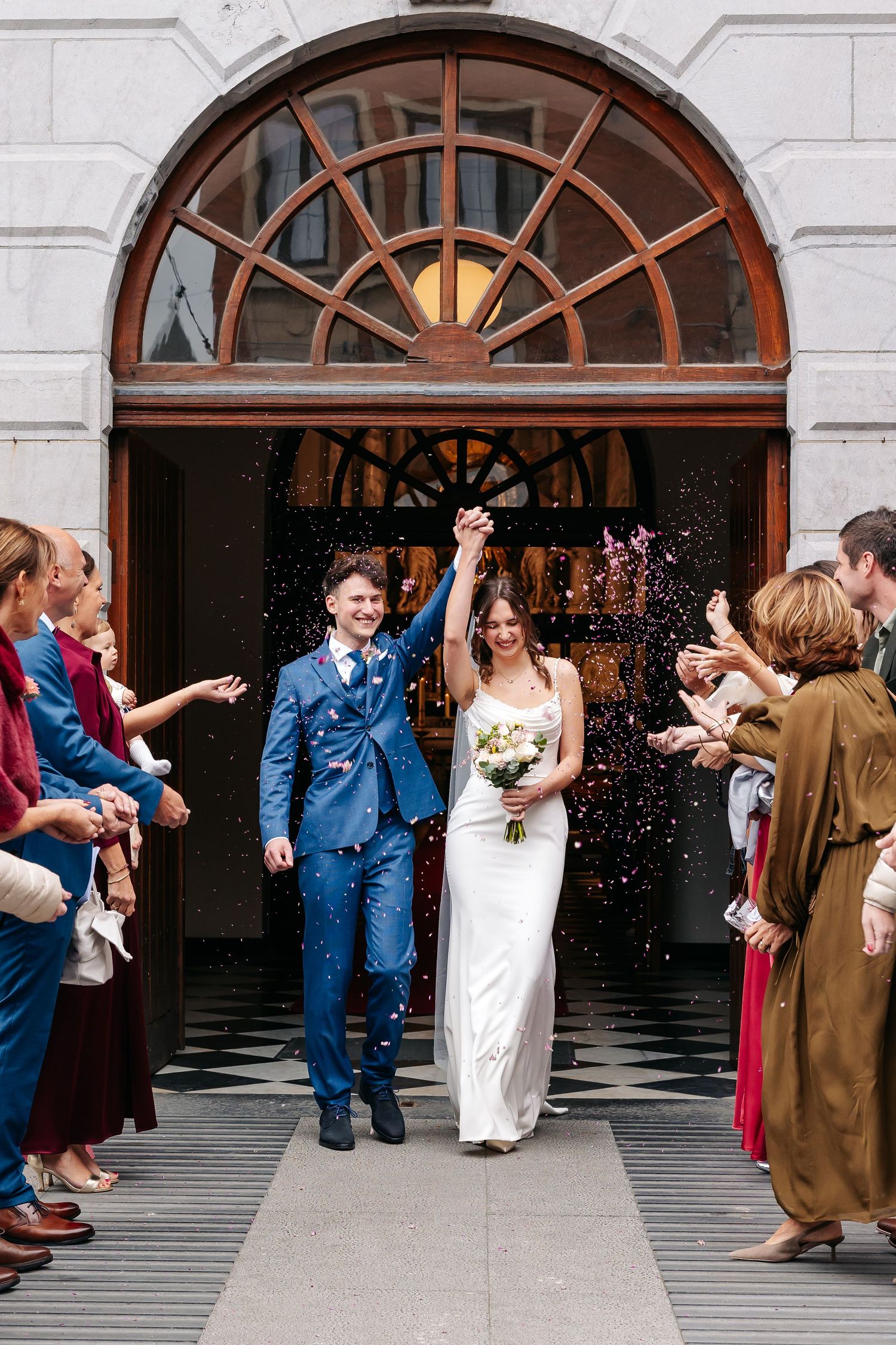 Smiling bride and groom exit building through confetti shower with guests.