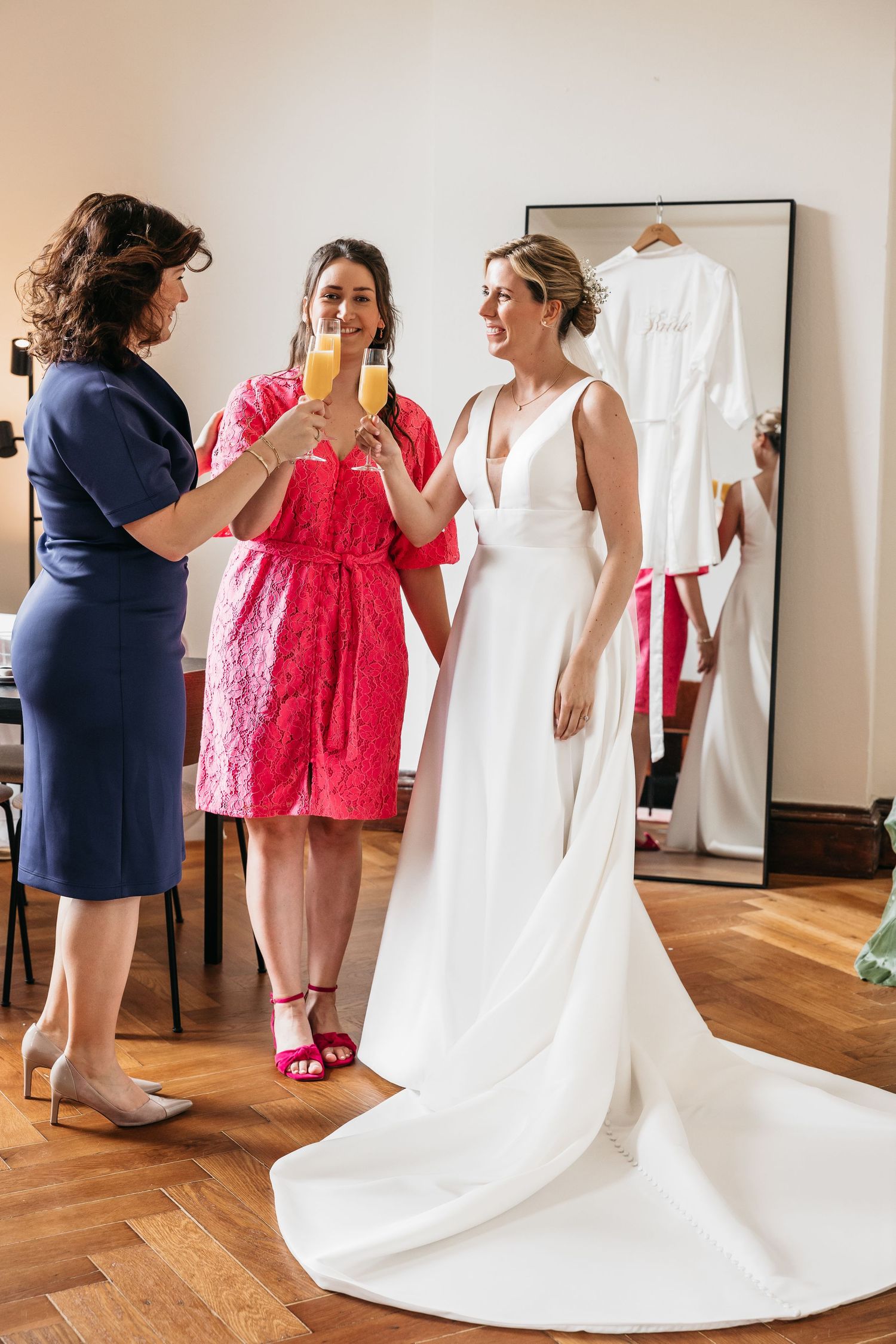 Bride and two bridesmaids toasting with mimosas in bright room with wedding robe on mirror.