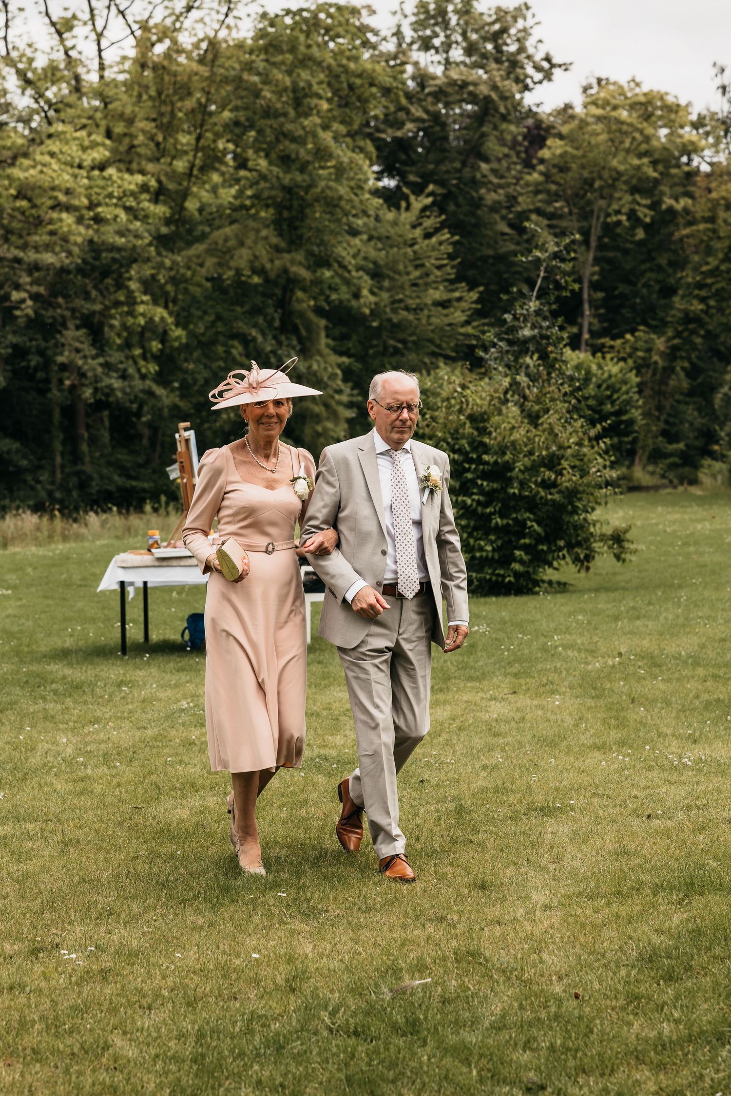 Elegant older couple walking arm in arm outdoors, woman in pink dress with hat, man in beige suit.