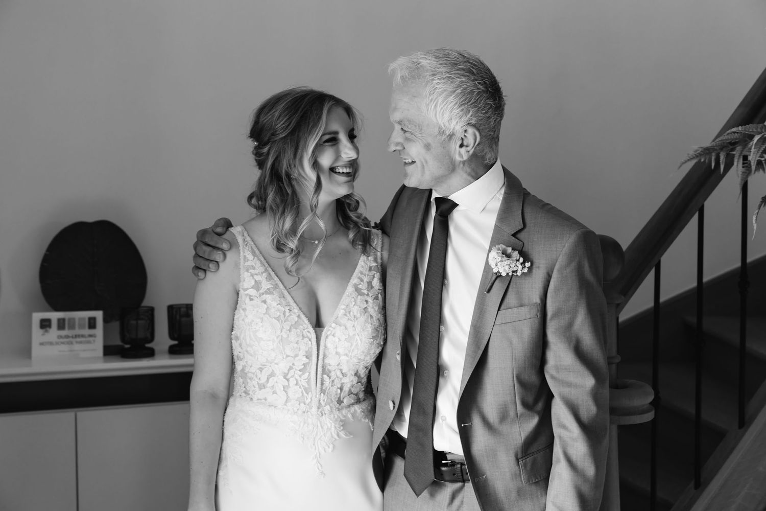 Black and white photo of bride and older man in suit laughing together near wooden staircase.