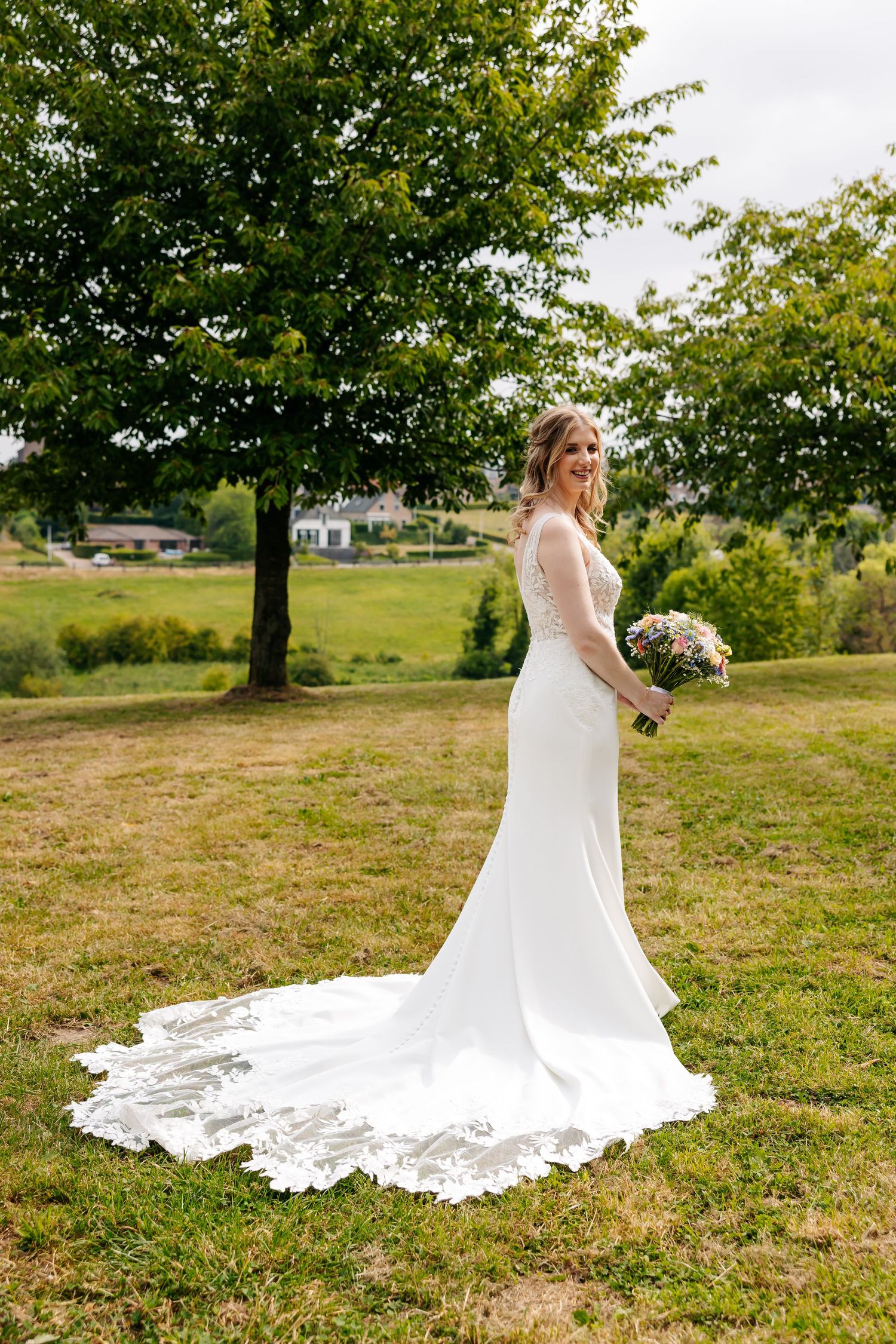 Bride in a fitted lace wedding gown with a long train standing on a green lawn.