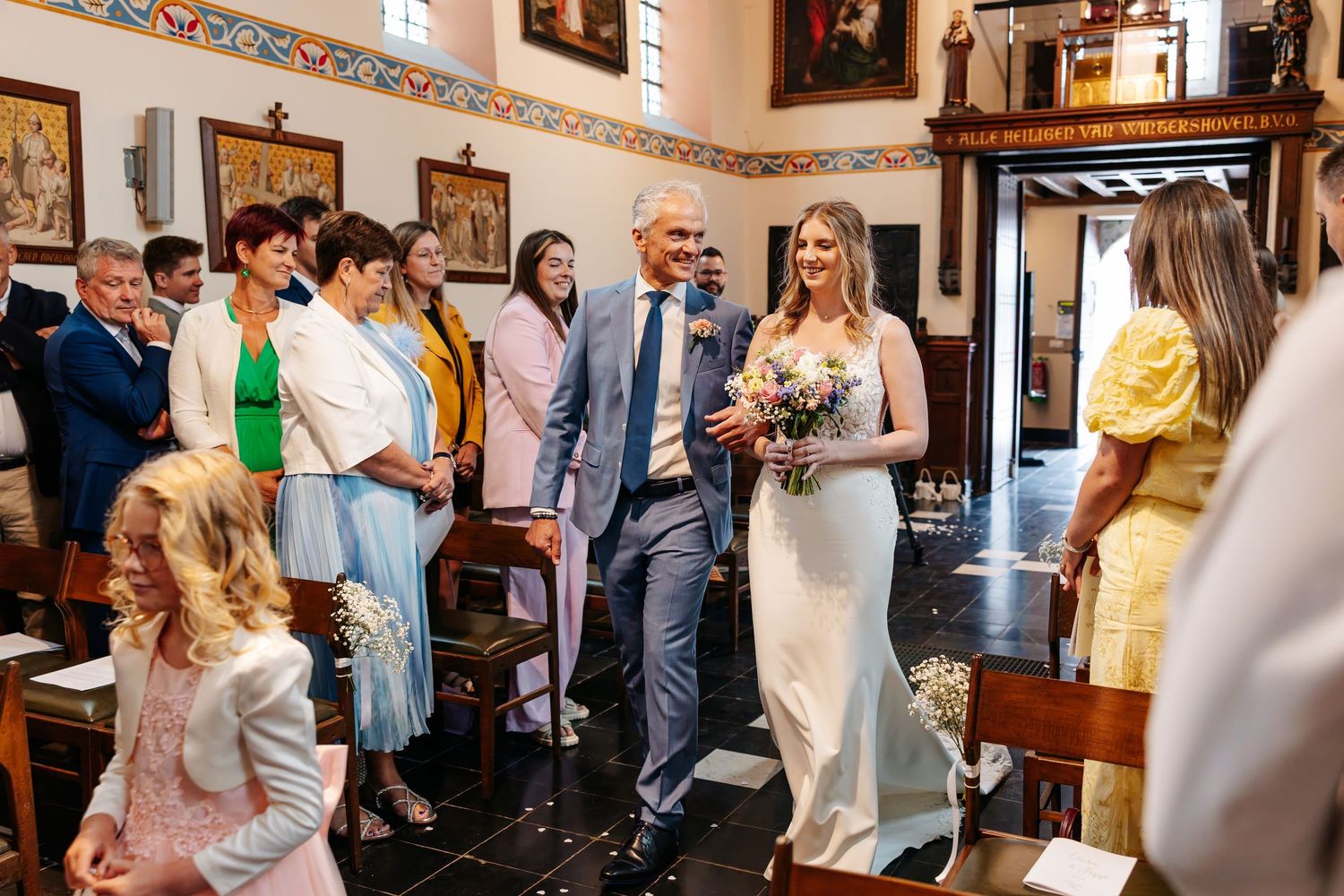 Bride and escort walk down decorated church aisle past smiling wedding guests.