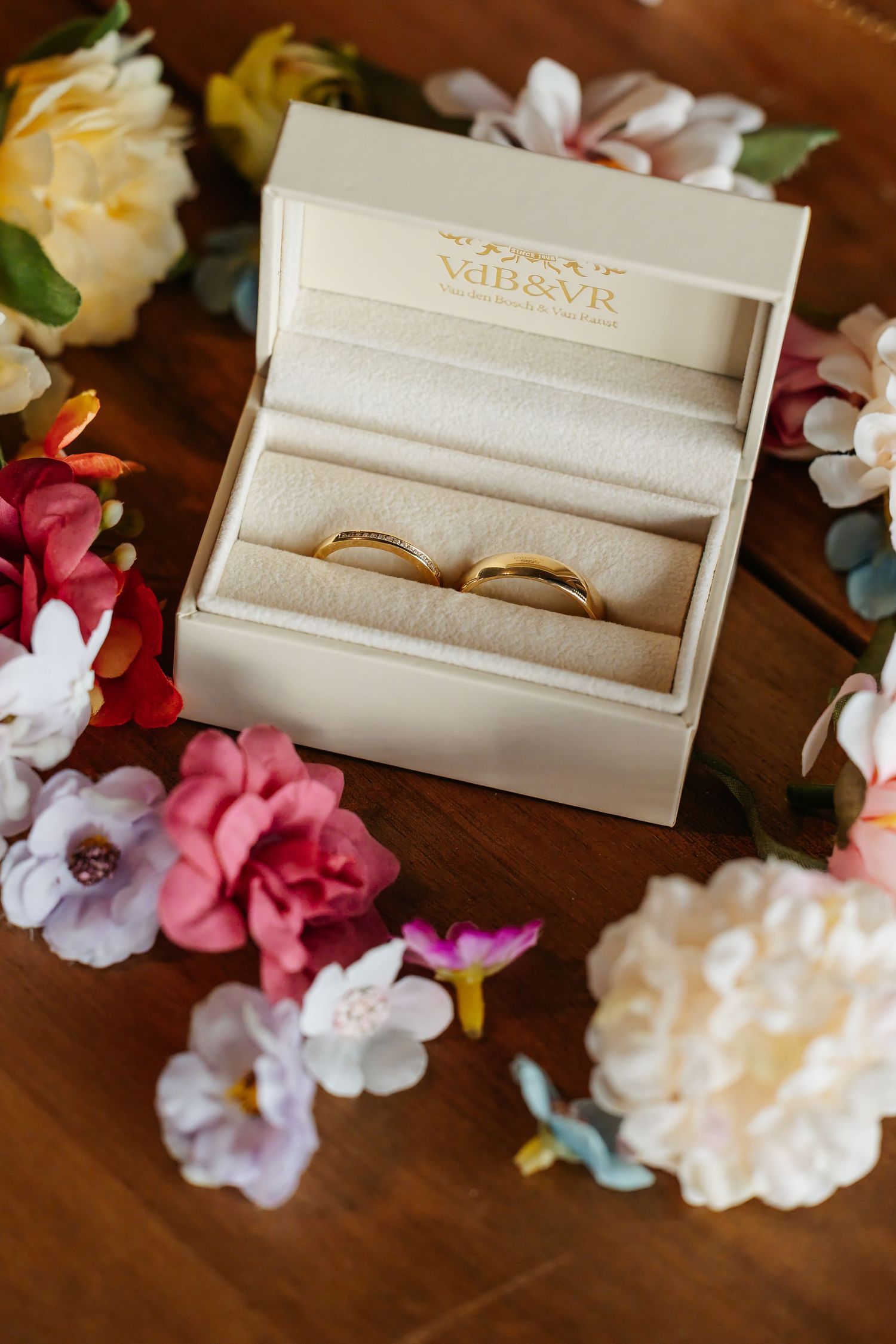 Two gold wedding bands displayed in a cream VdB&VR jewelry box surrounded by colorful flowers.