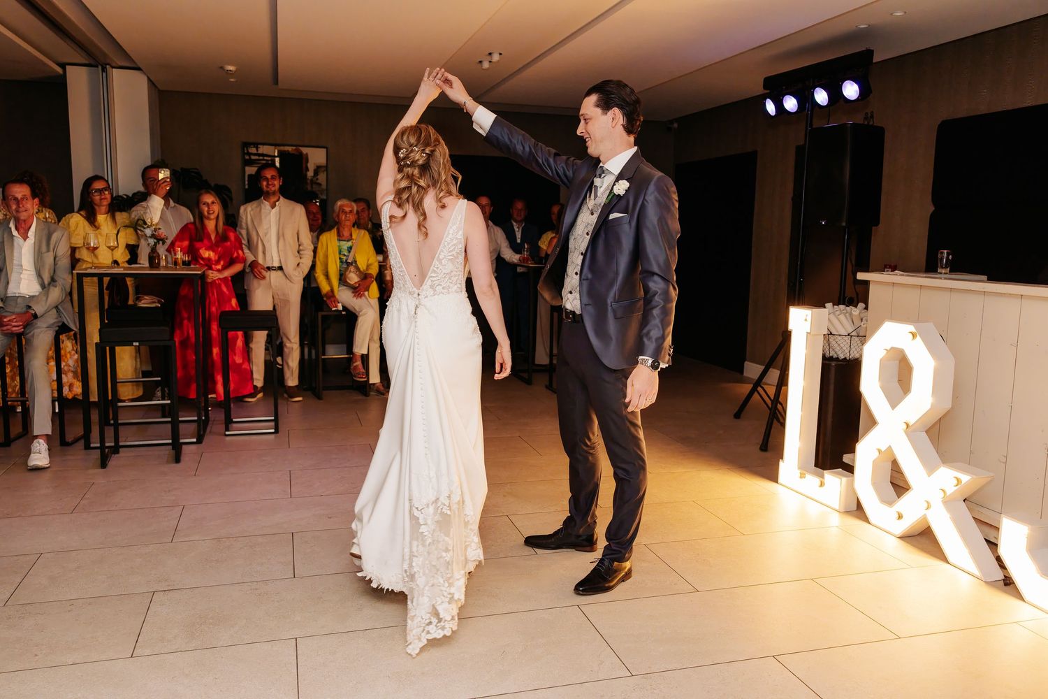Groom twirls bride in elegant wedding dress during first dance with illuminated letter decorations.