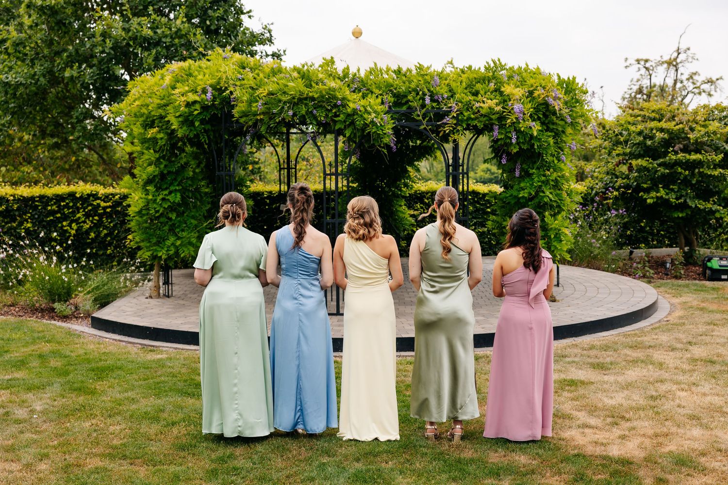 Five bridesmaids in pastel dresses viewed from behind facing a garden gazebo at wedding.