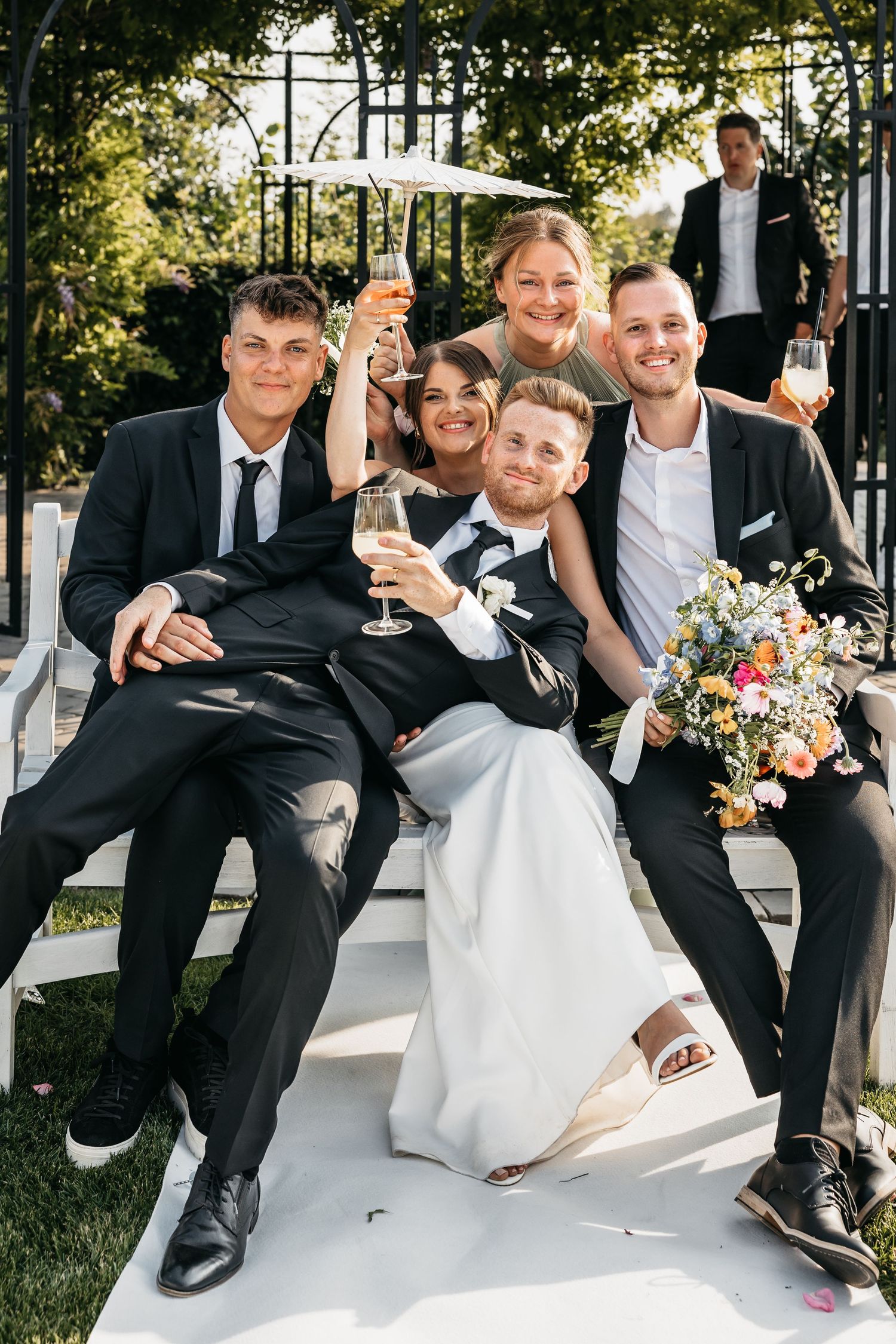 Bride and groom sit with wedding guests on white steps toasting with champagne glasses.