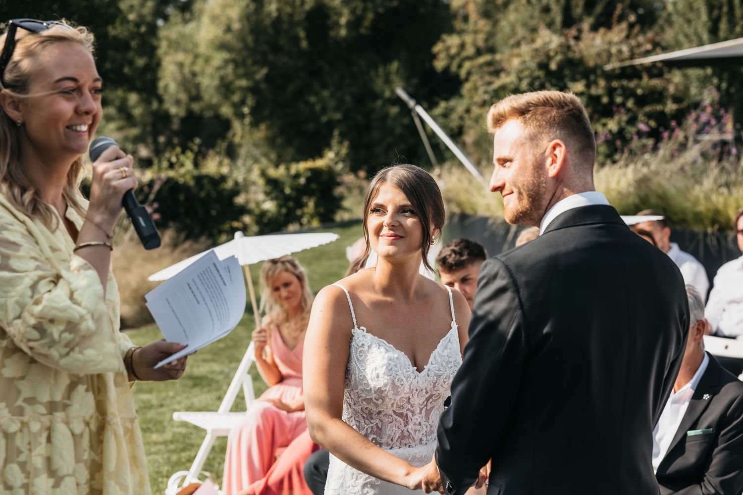 Bride and groom hold hands smiling during sunny outdoor wedding ceremony with officiant.