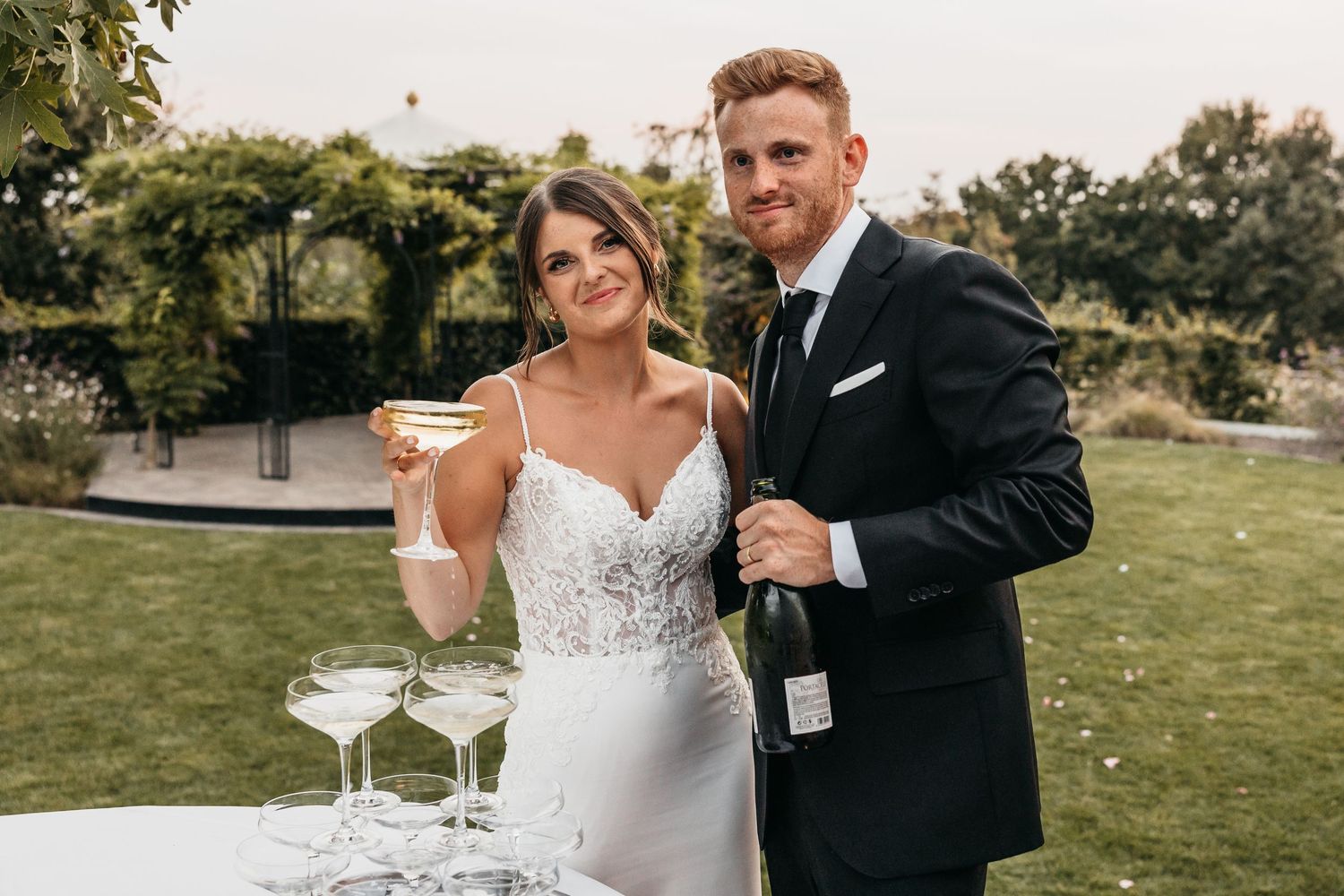 Smiling bride and groom posing with champagne bottle and glasses at outdoor wedding reception.