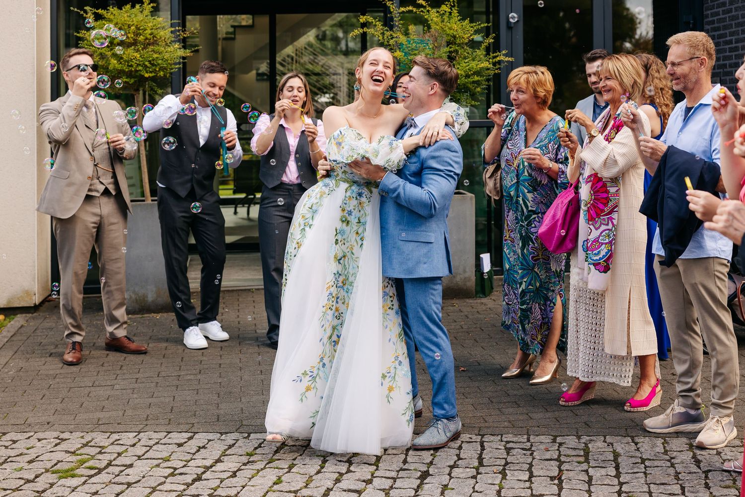 Bride in floral gown laughs joyfully with groom as wedding guests celebrate around them.