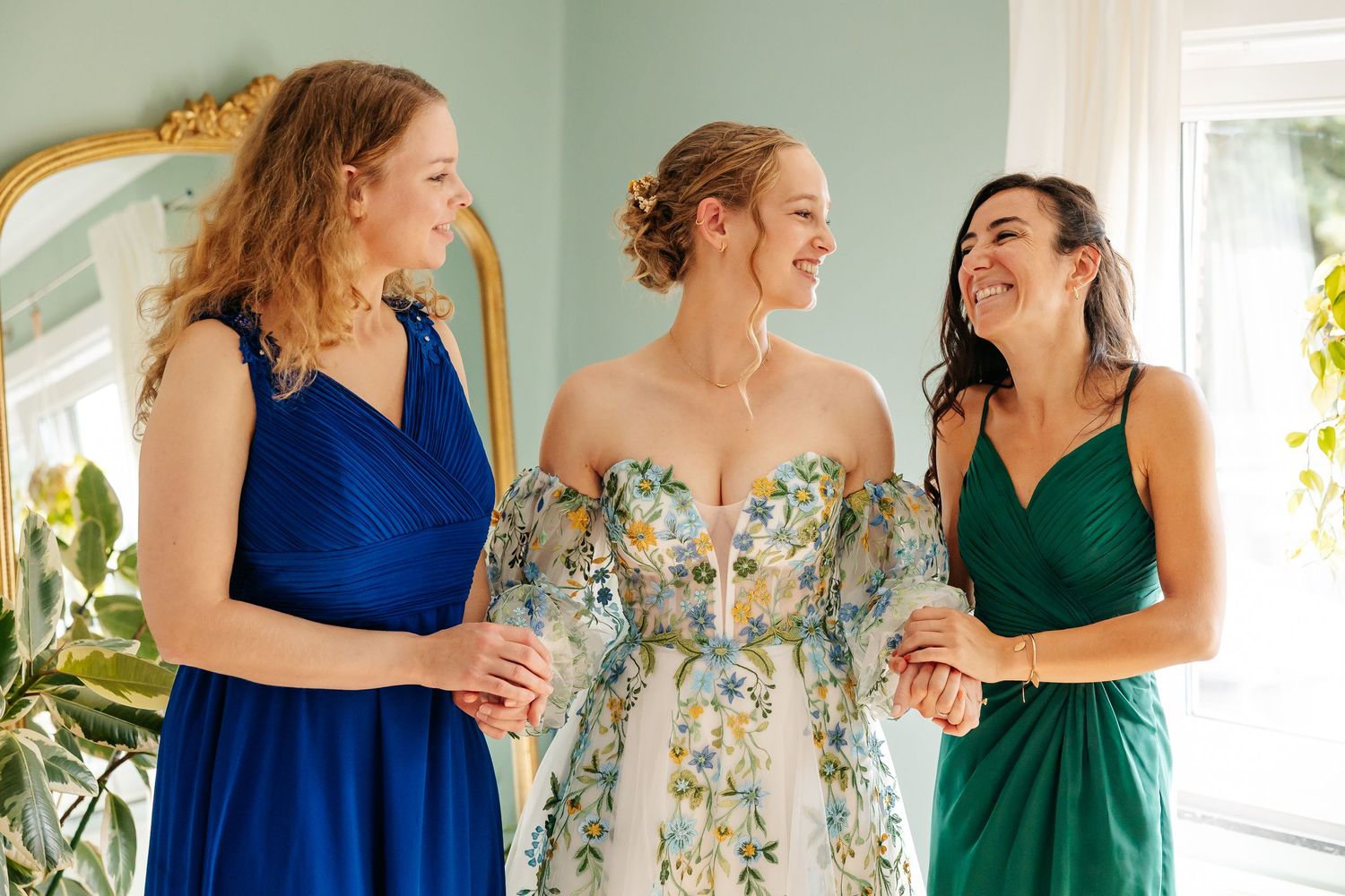 Bride in floral gown smiling and holding hands with two bridesmaids in blue and green.