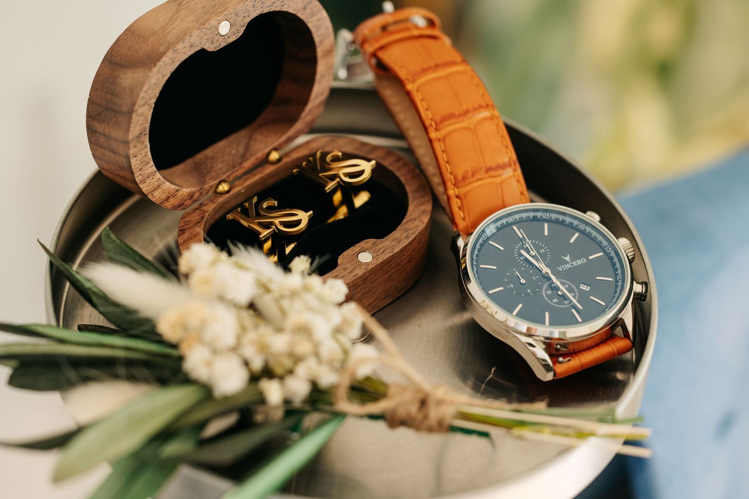 Brown leather watch beside a wooden ring box and white flowers on a silver tray.
