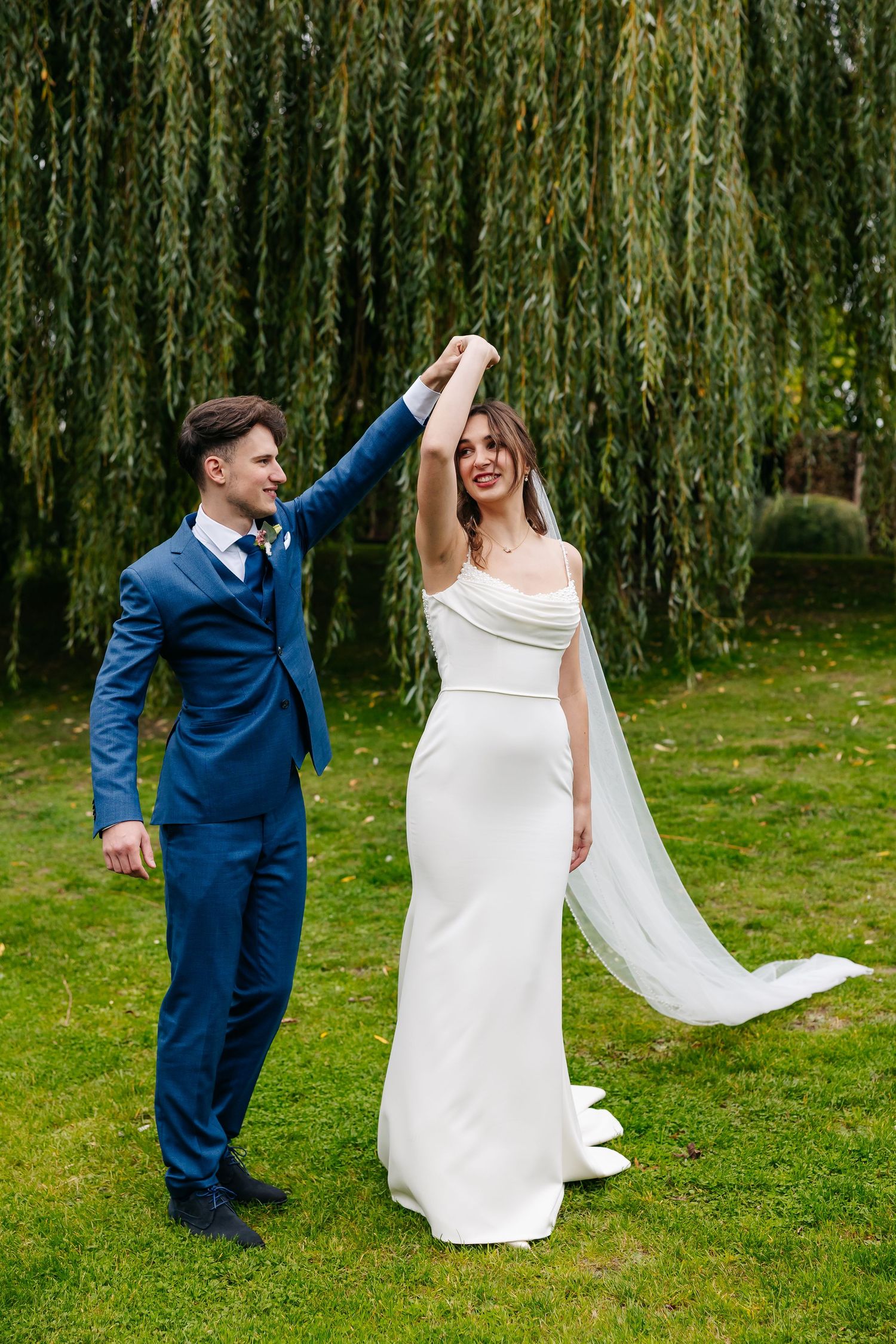 Groom spinning bride in elegant white gown on green lawn beneath a weeping willow.