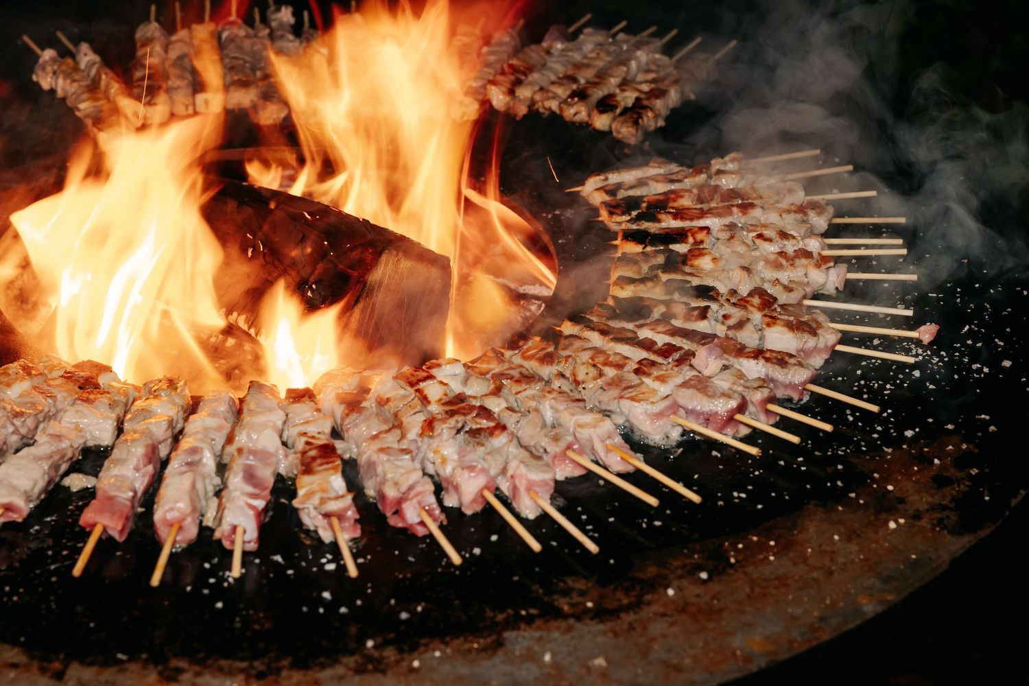 Meat skewers grilling over open flame on large circular barbecue grill at outdoor event.