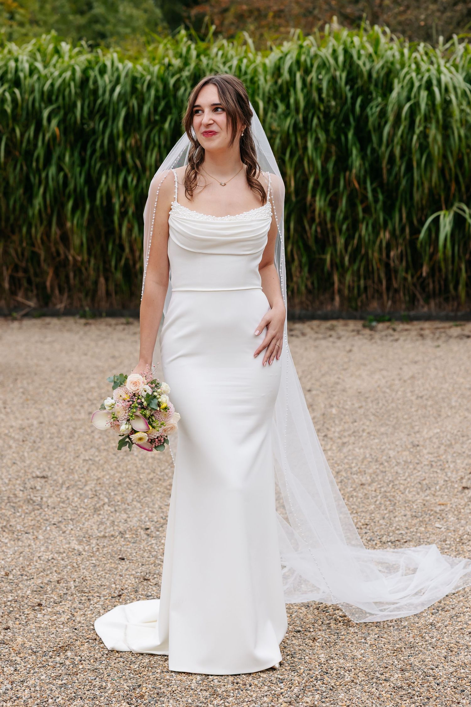 Elegant bride in fitted white gown and veil holding pastel bouquet outdoors near green foliage.