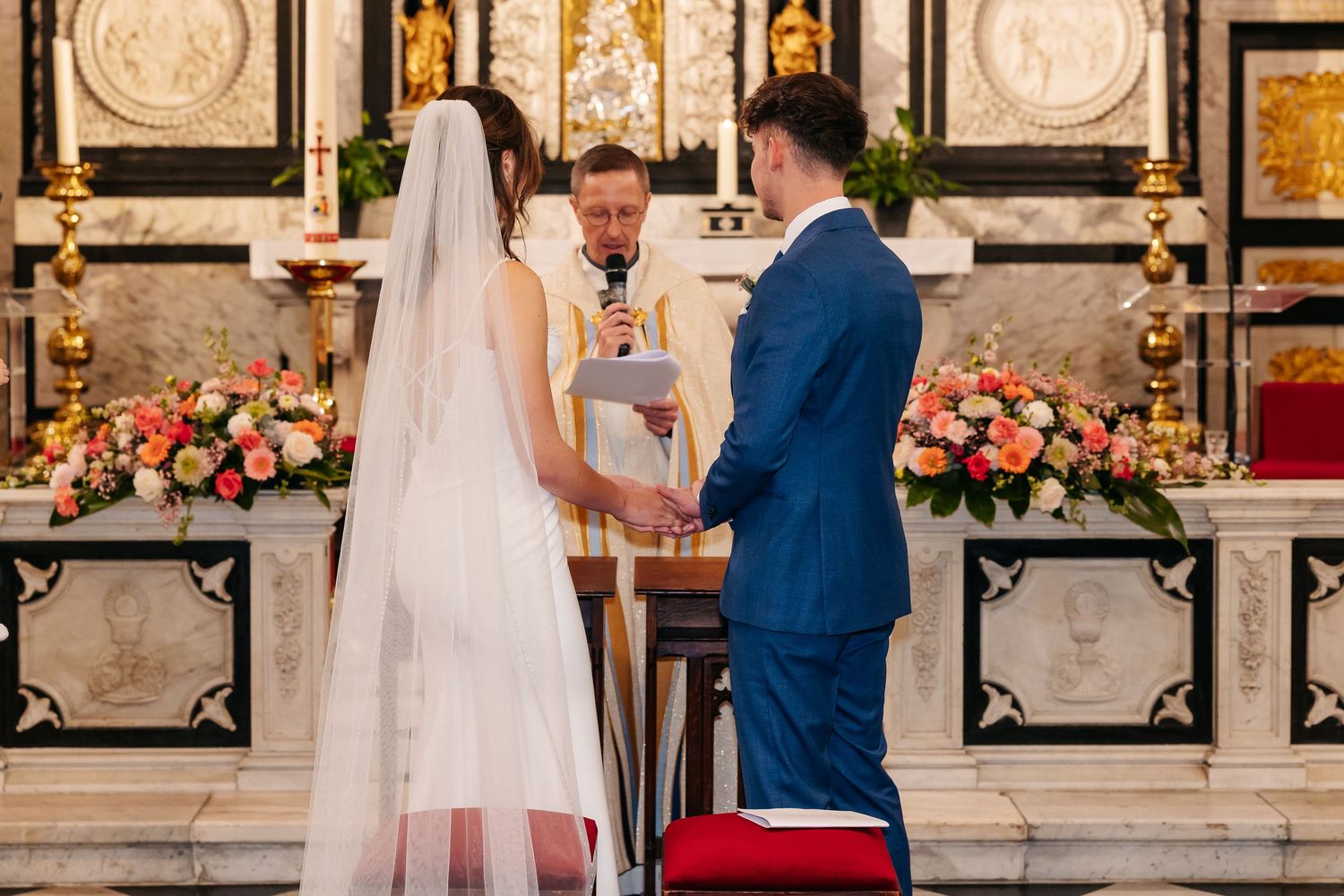 Priest officiates wedding ceremony as bride and groom hold hands before floral altar.