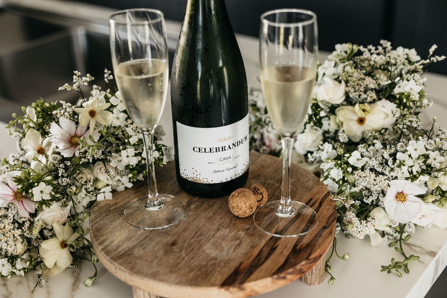 Celebrand champagne bottle with two filled flutes on wooden board surrounded by white wedding flowers.