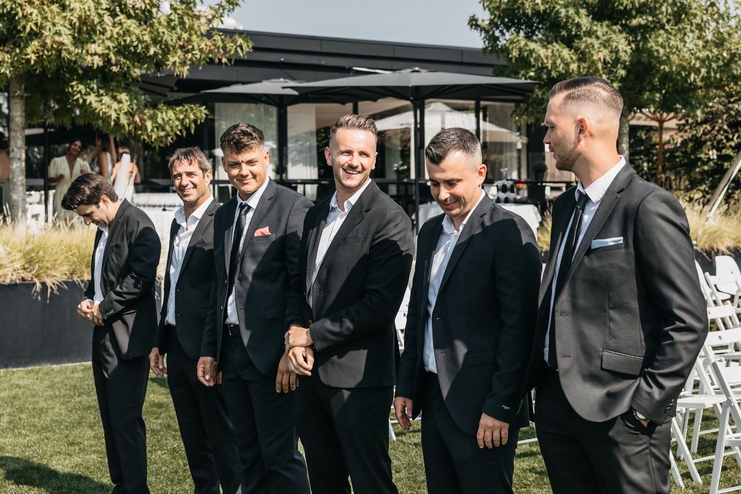 Five groomsmen in dark suits laughing together beside white chairs at outdoor wedding venue.