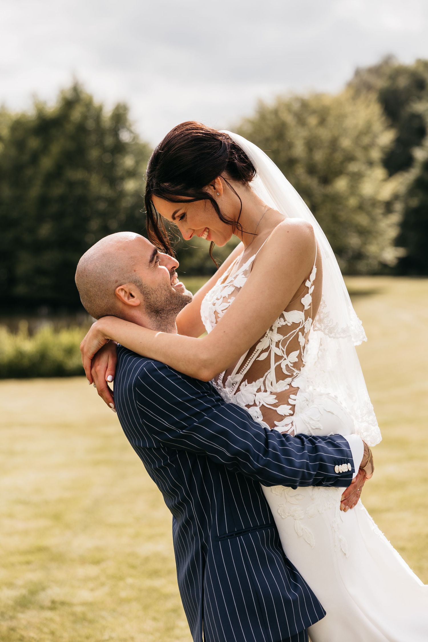Groom lifts smiling bride in lace wedding dress outdoors with lush green trees in background.