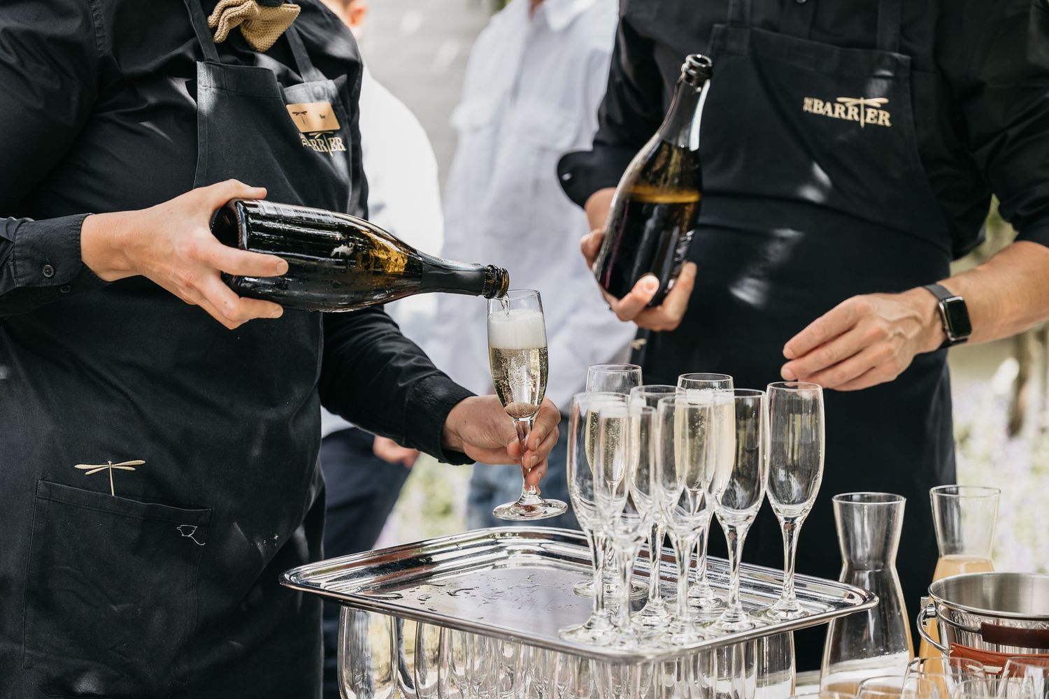 Two servers in black aprons pouring champagne into crystal flutes arranged on a silver tray.