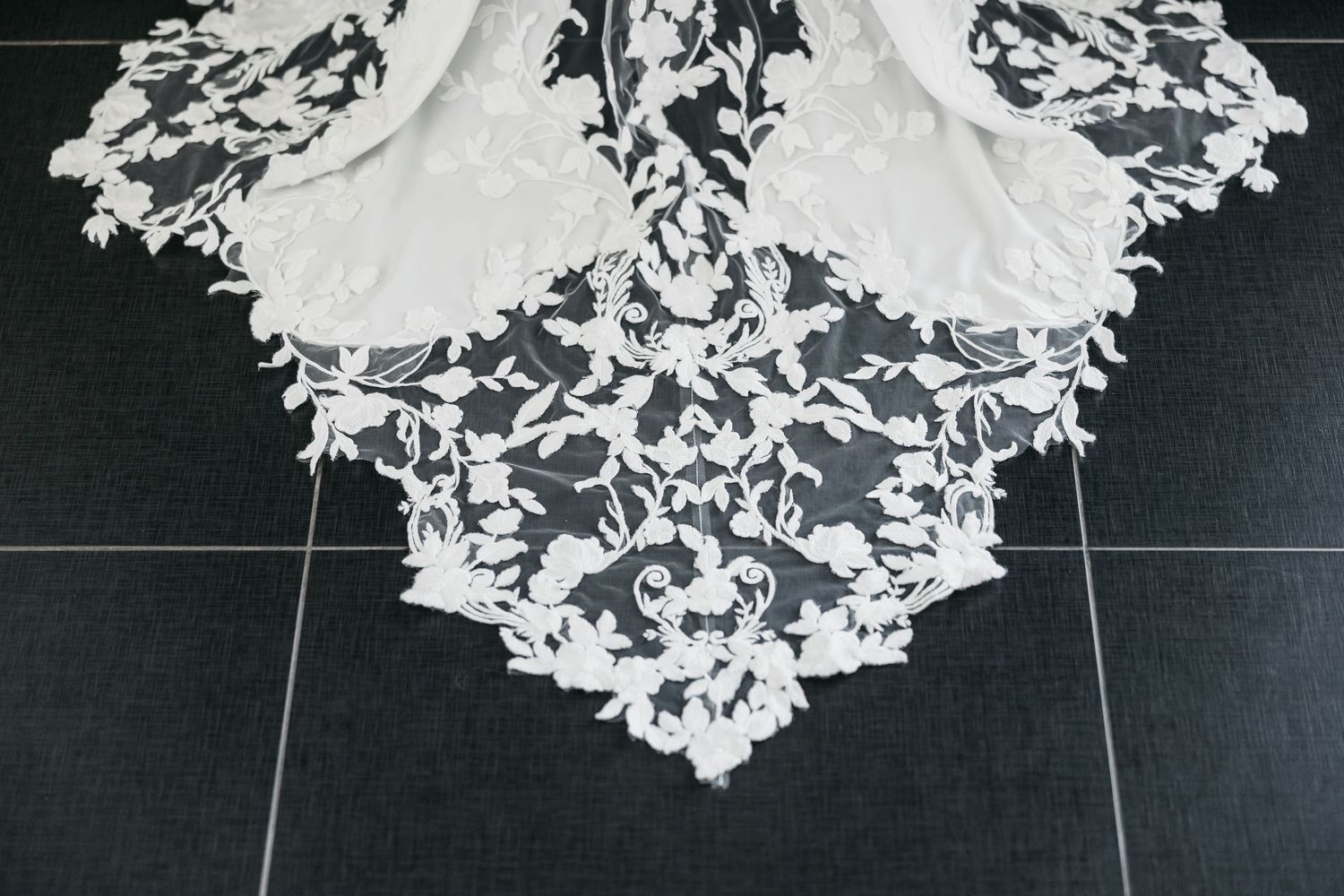 Intricate white floral lace wedding dress train spread on dark tile floor.