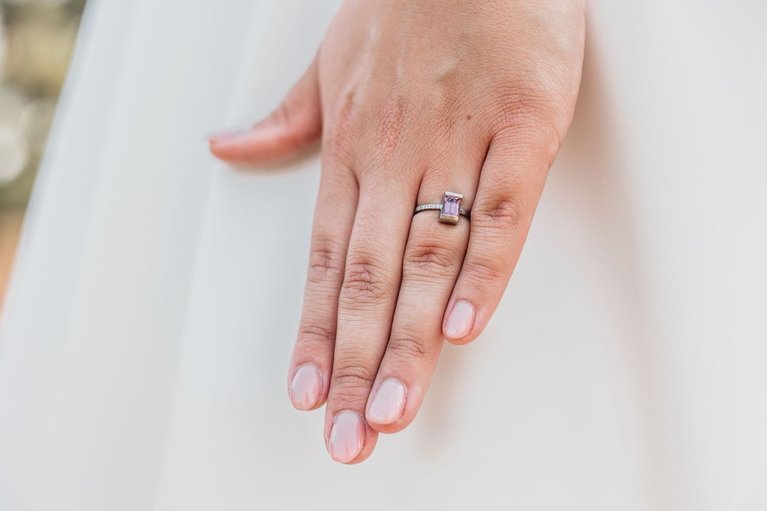 Close-up of bride's hand displaying elegant square-cut diamond engagement ring on white dress.