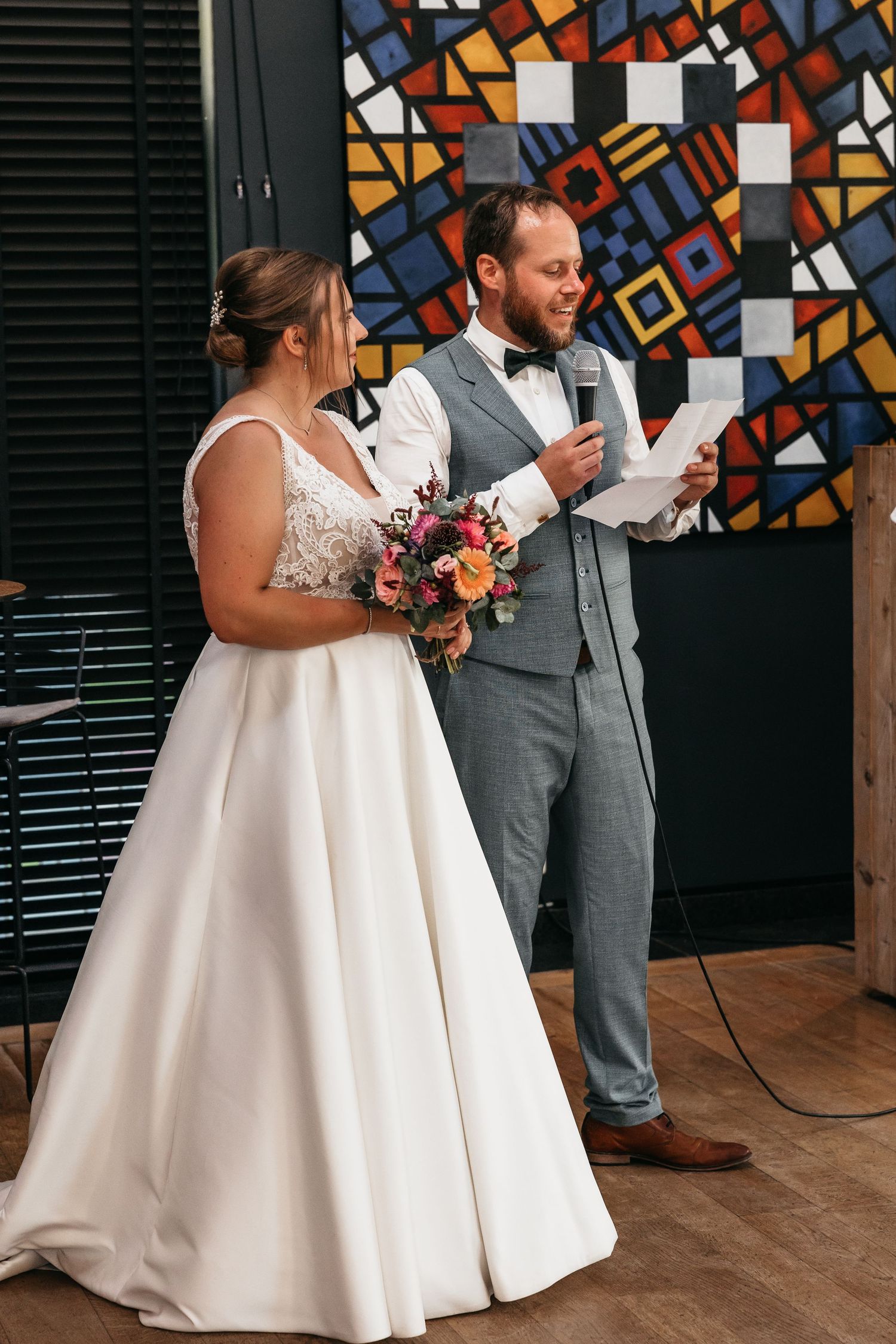Bride holding colorful bouquet stands beside groom in gray suit reading vows at wedding ceremony.