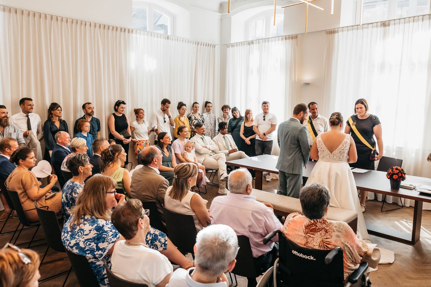 Wedding ceremony taking place in a bright room with guests seated and standing around the couple.