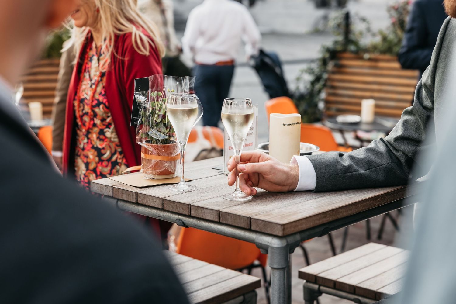 Wedding guests enjoying champagne at outdoor cafe table in elegant attire on sunny day.