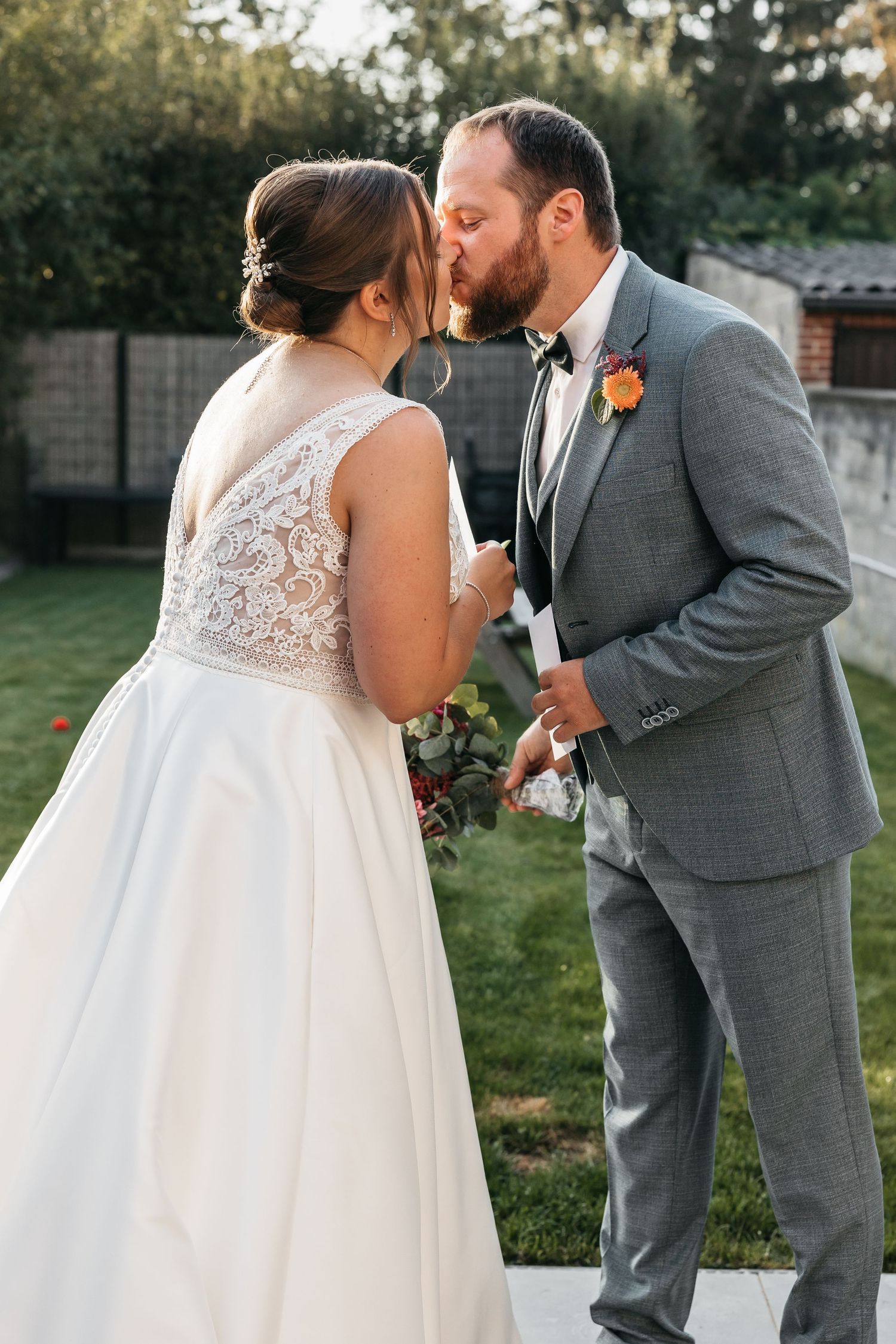 Groom in gray suit and bow tie kisses bride in lace gown tenderly outdoors after vow exchange.