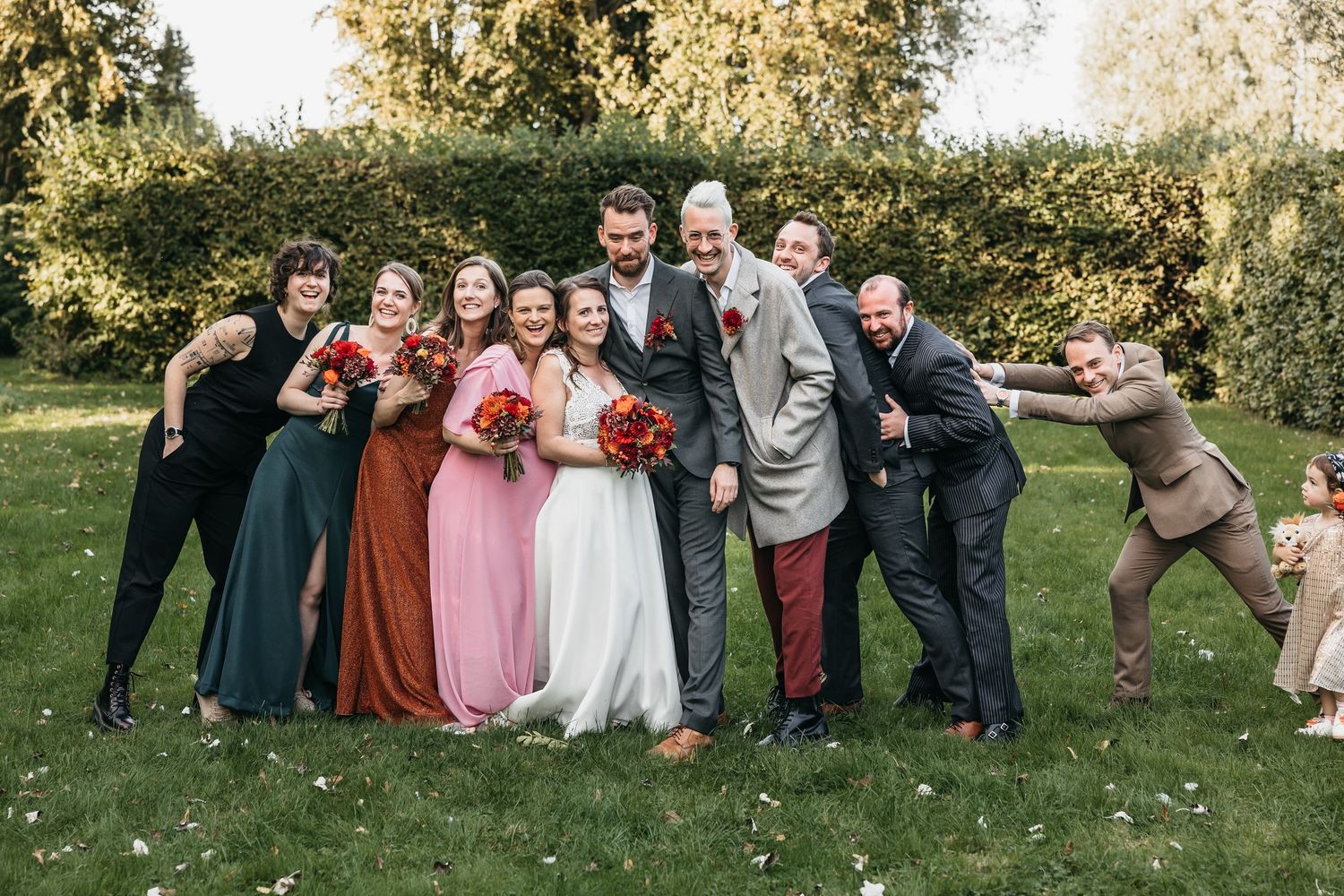 Wedding party posing playfully on green lawn with bride in white gown and bridesmaids in colorful dresses holding red bouquets.