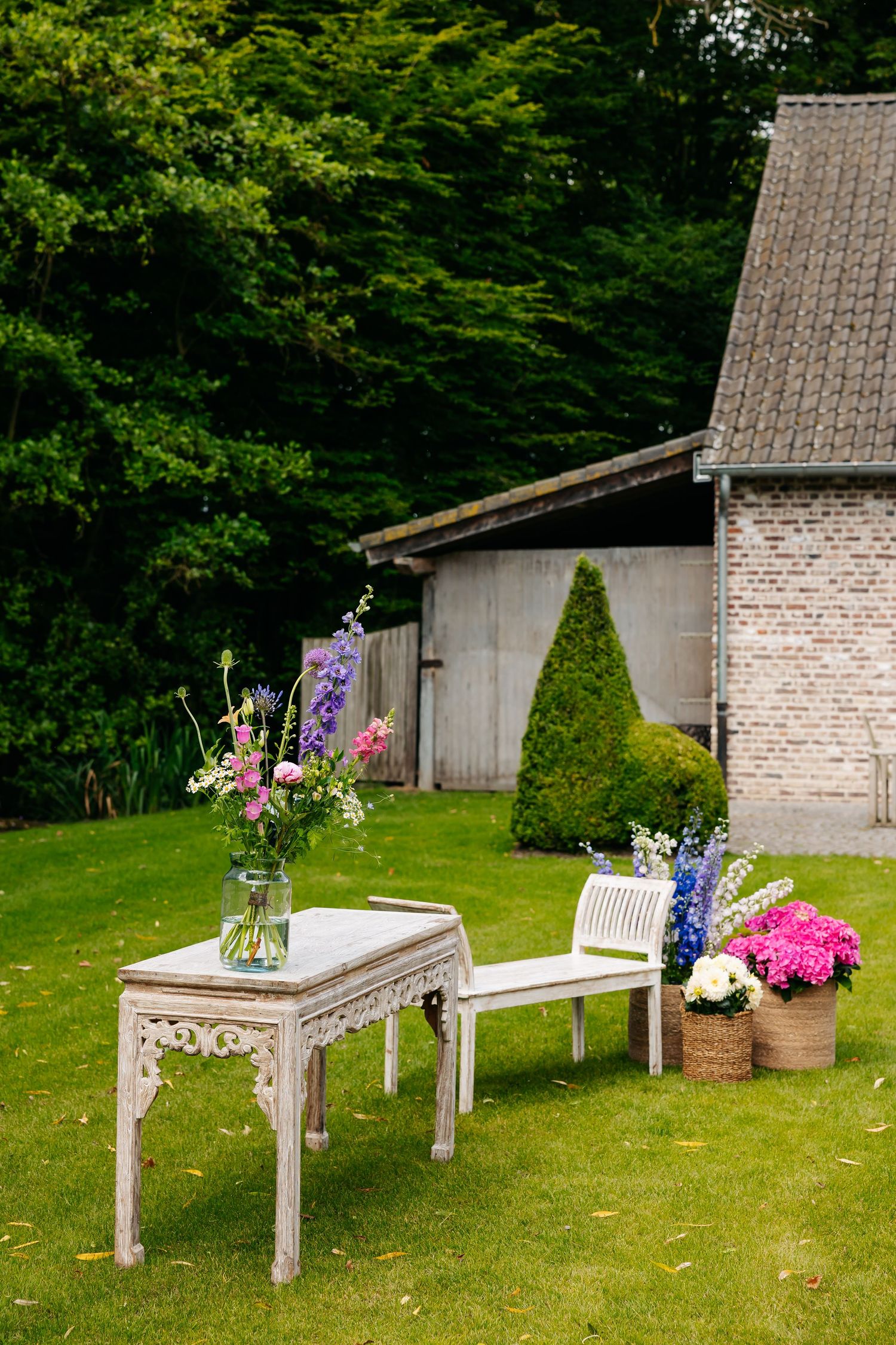 Elegant outdoor wedding setup with ornate white table, floral arrangement, bench, and potted flowers on green lawn.