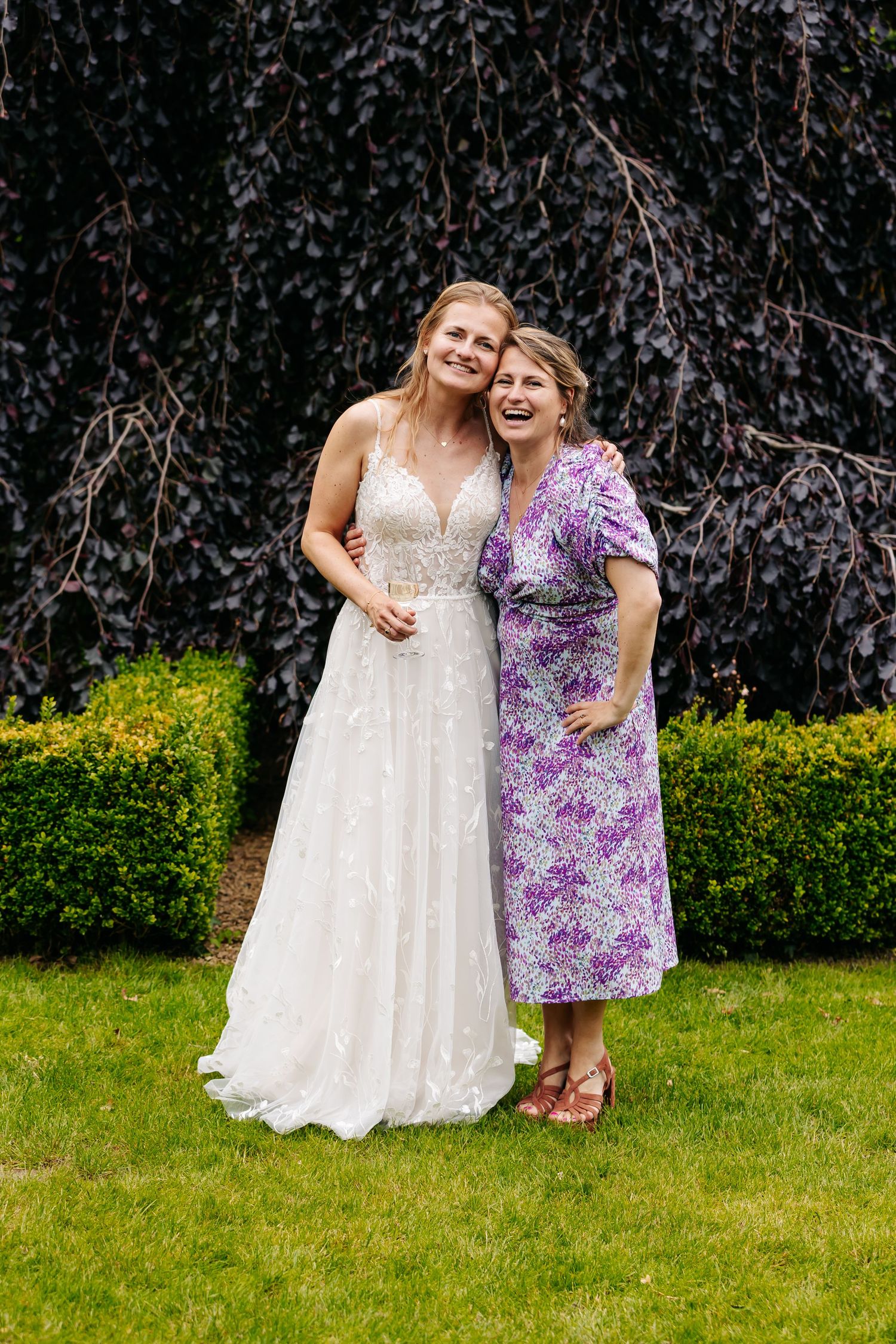 A smiling bride in a white lace gown poses with an older woman in a purple floral dress.