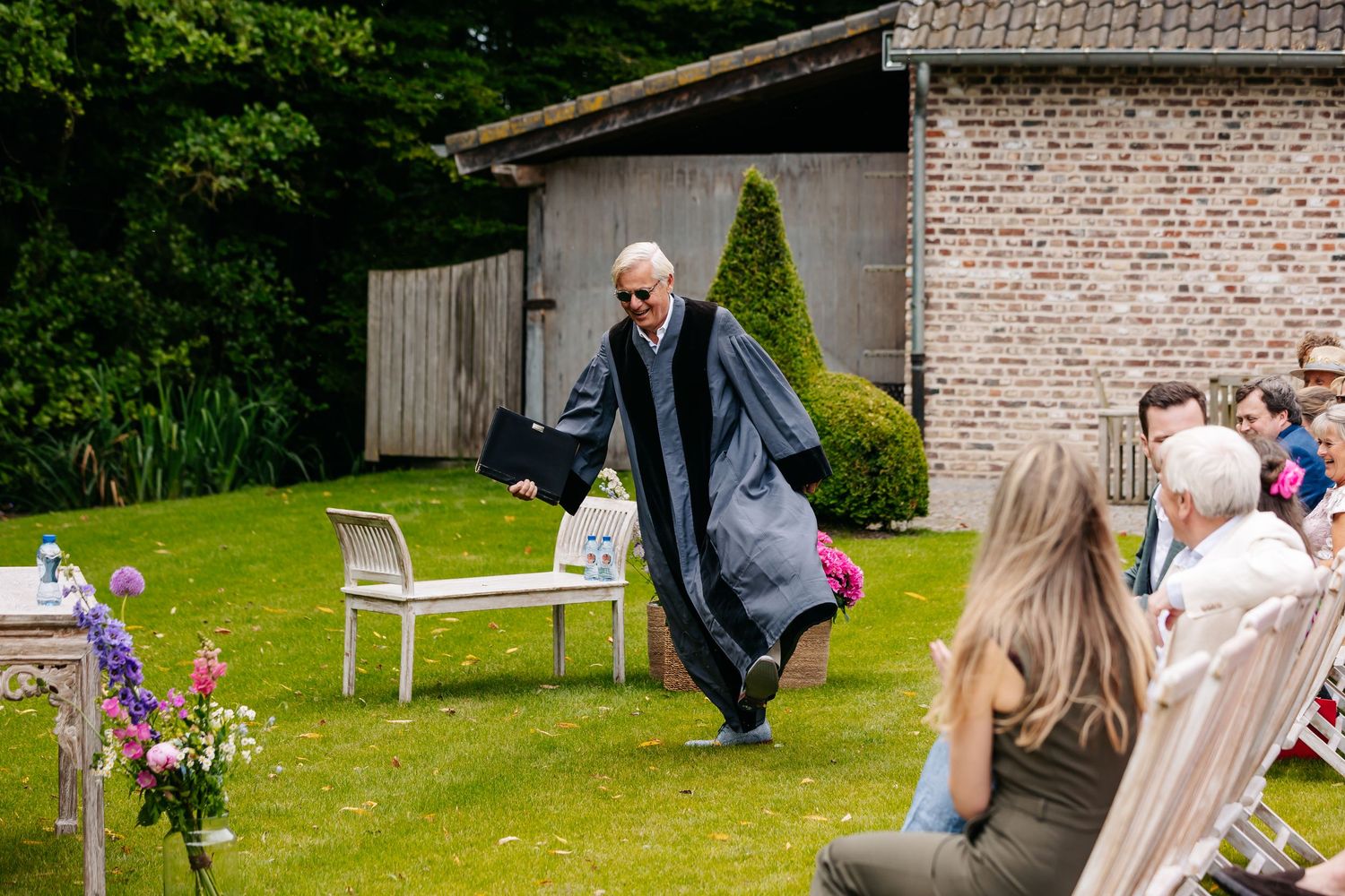 Officiant in a gray robe walking toward guests at an outdoor garden wedding ceremony.