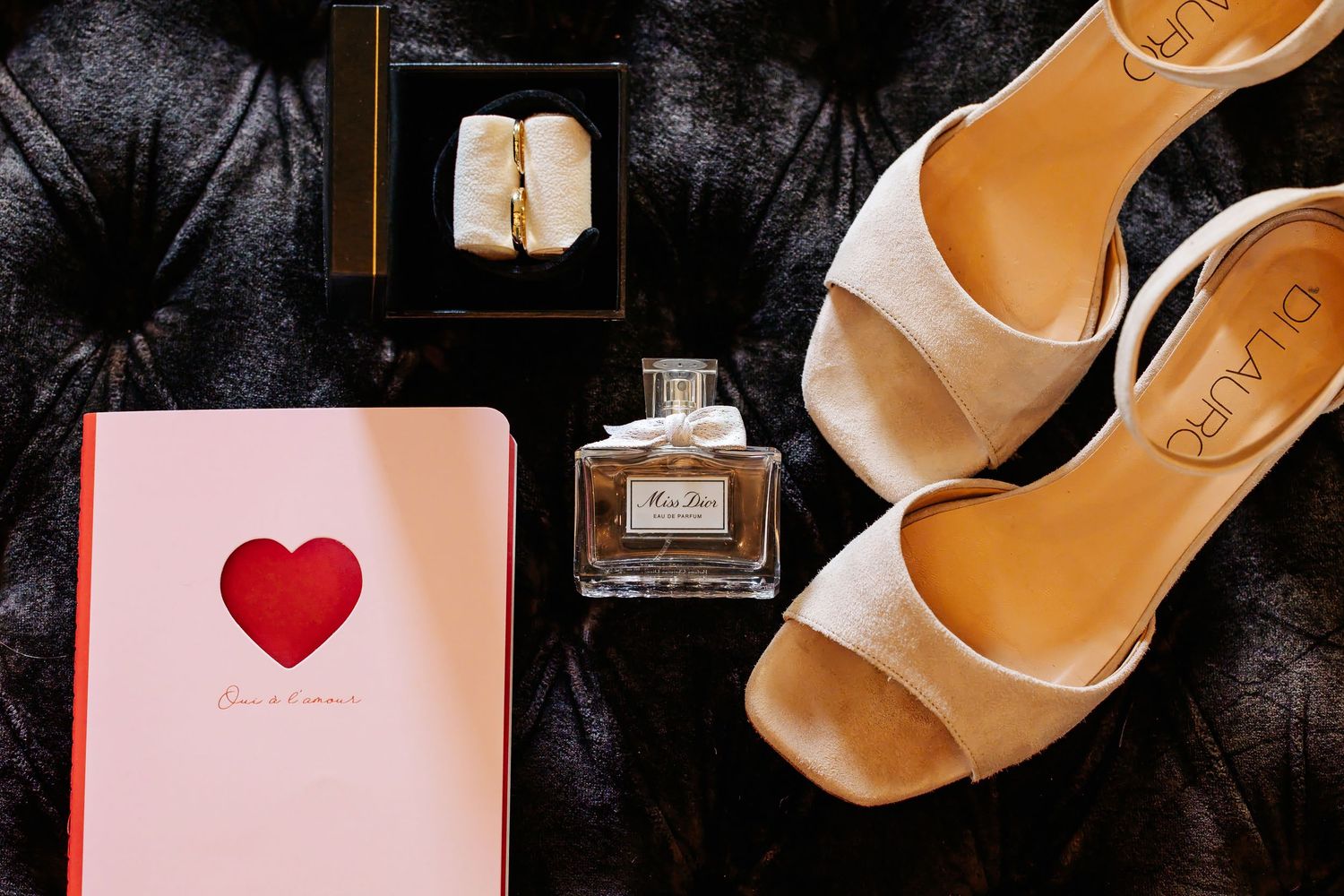 Wedding flat lay with heels, perfume, ring box, and heart journal on dark velvet.