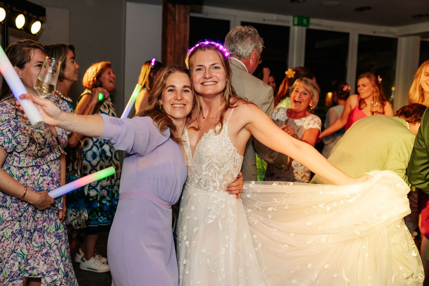 Bride in white lace gown dancing with smiling guest holding glow stick at lively wedding reception party.