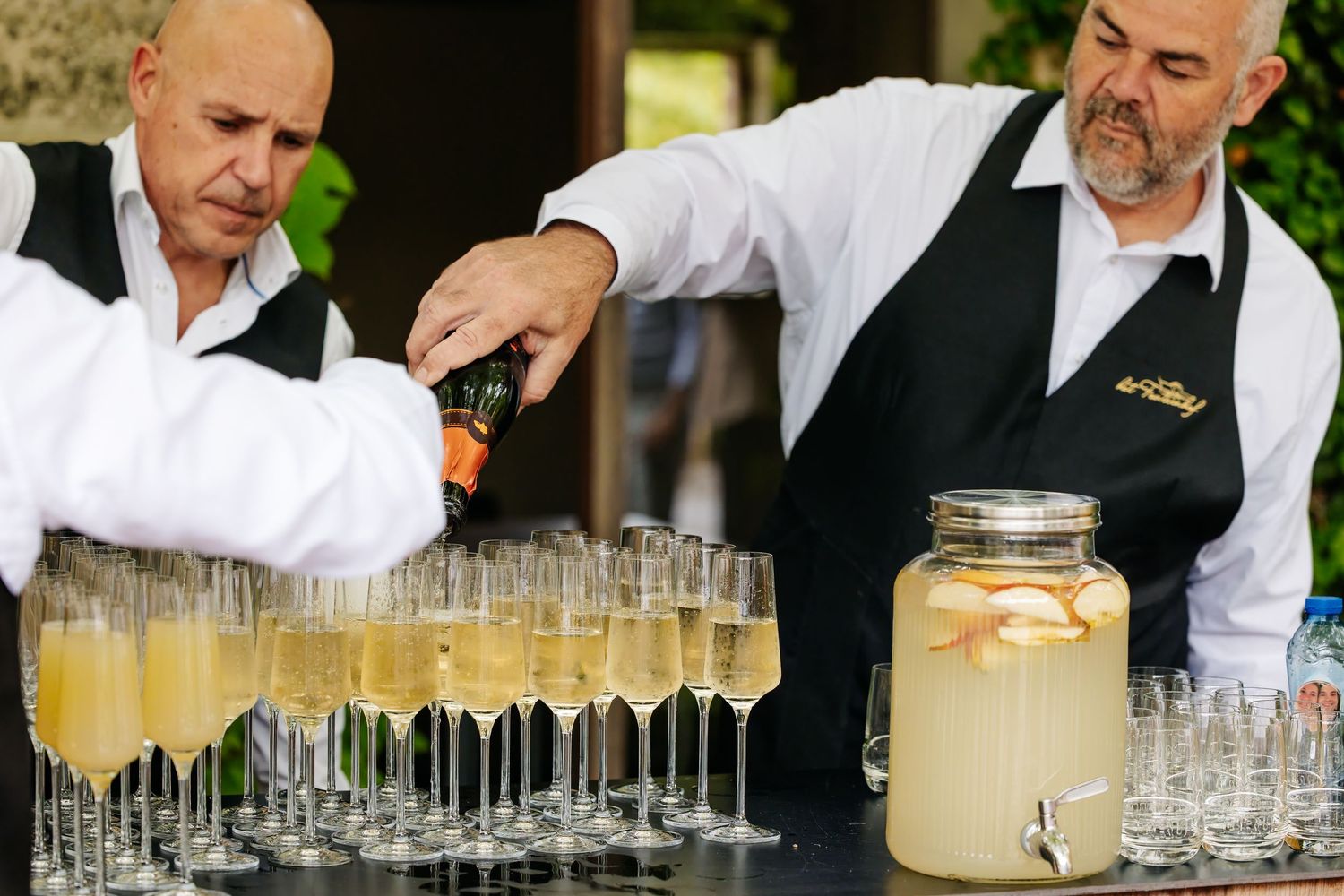 Two bartenders in black aprons pour champagne into elegant flute glasses at a wedding reception bar.
