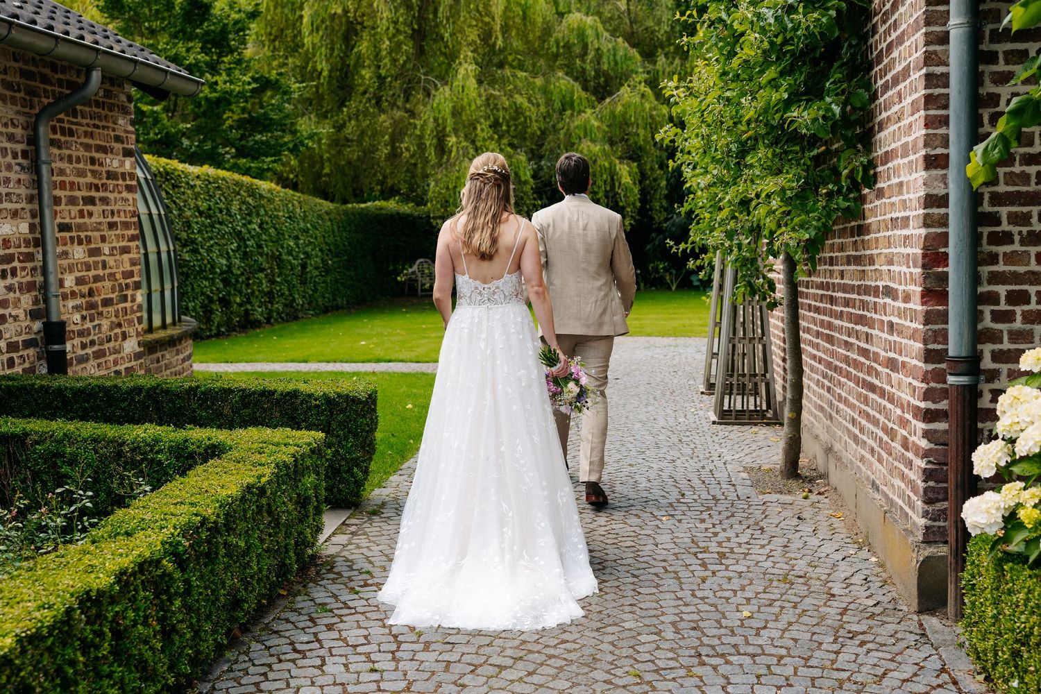 Bride and groom walking away along a cobblestone path lined with manicured hedges and brick walls.