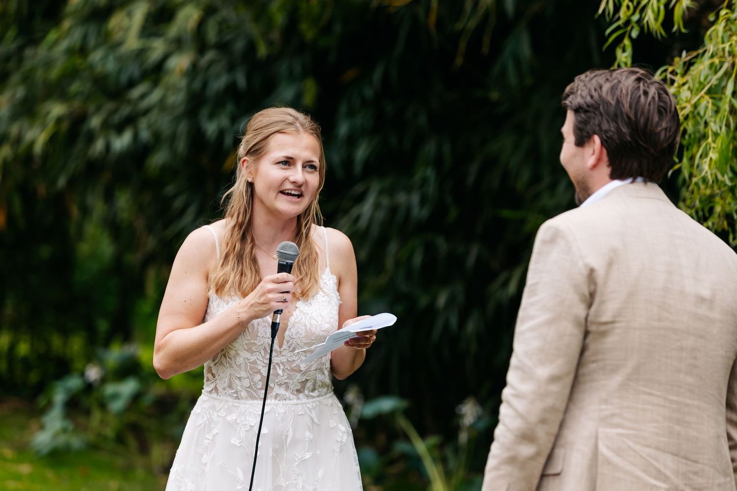 Bride in lace wedding dress speaking heartfelt vows into microphone during outdoor garden ceremony.