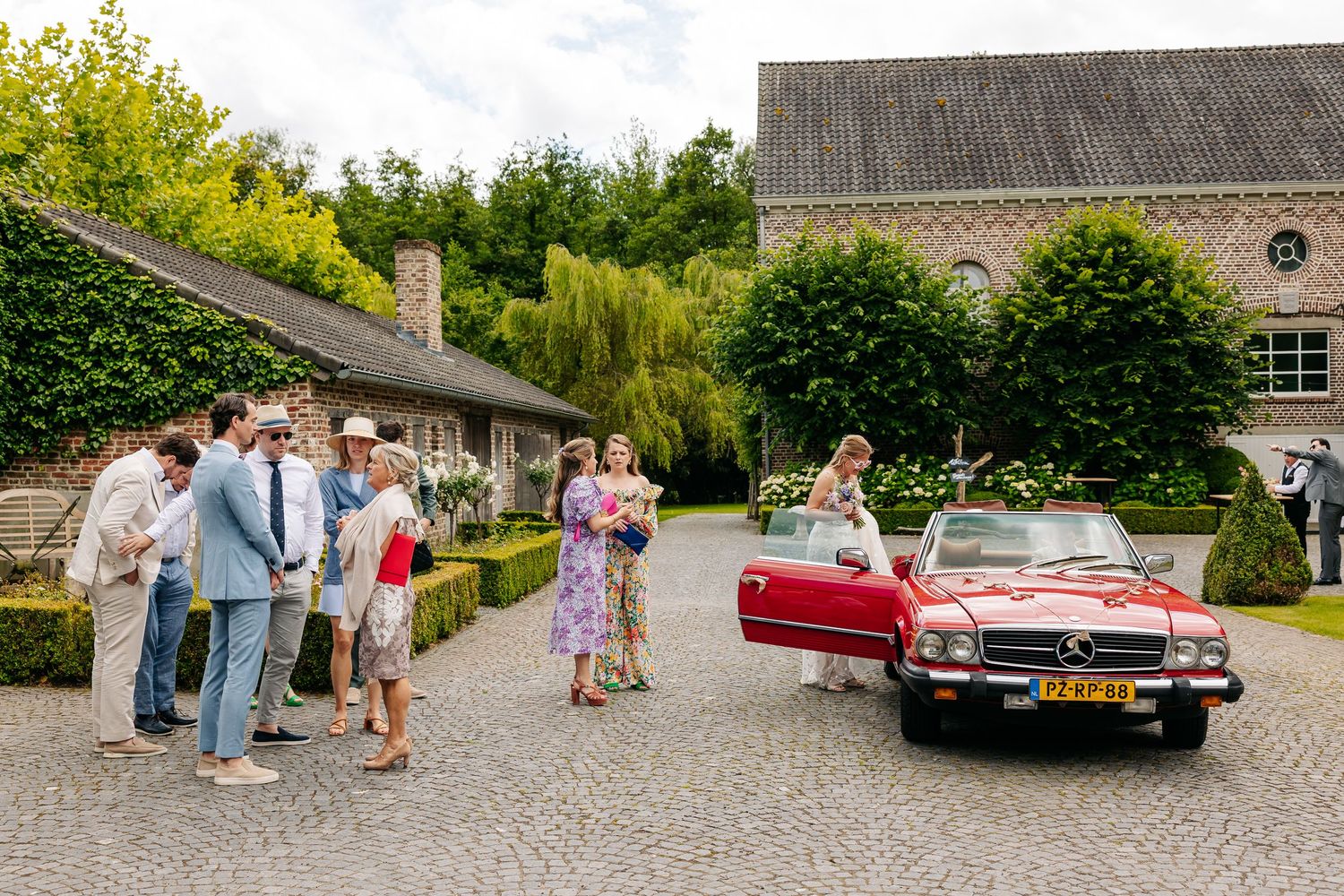 Wedding guests gathering around a classic red Mercedes convertible on a cobblestone courtyard.