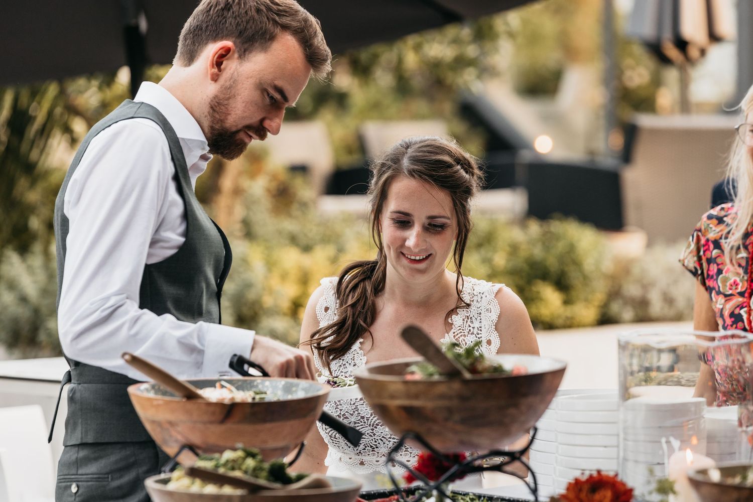 Smiling couple serving themselves from a rustic wooden salad bowl at an outdoor catered event.