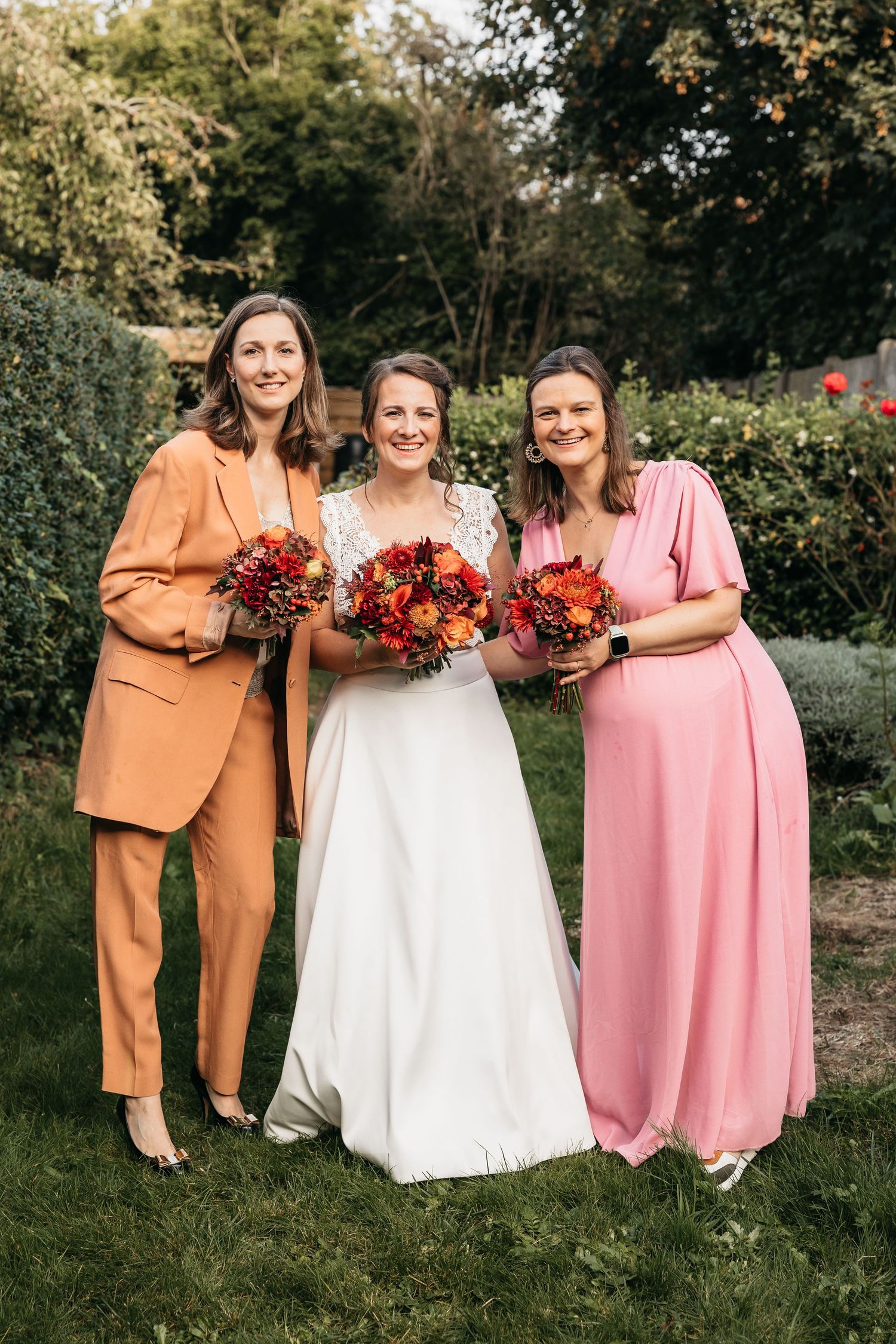 Bride in white gown with two bridesmaids in orange suit and pink dress holding autumn bouquets in garden.