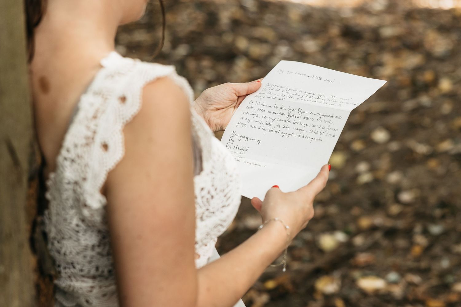 Bride in lace wedding dress reading a handwritten letter outdoors among autumn leaves.