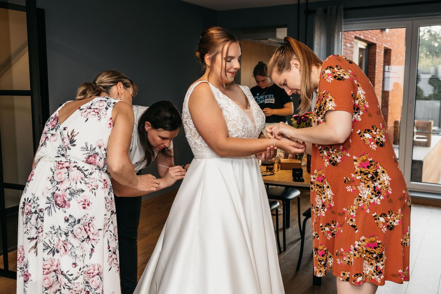 Bride in lace wedding dress surrounded by bridesmaids helping with final preparations indoors.