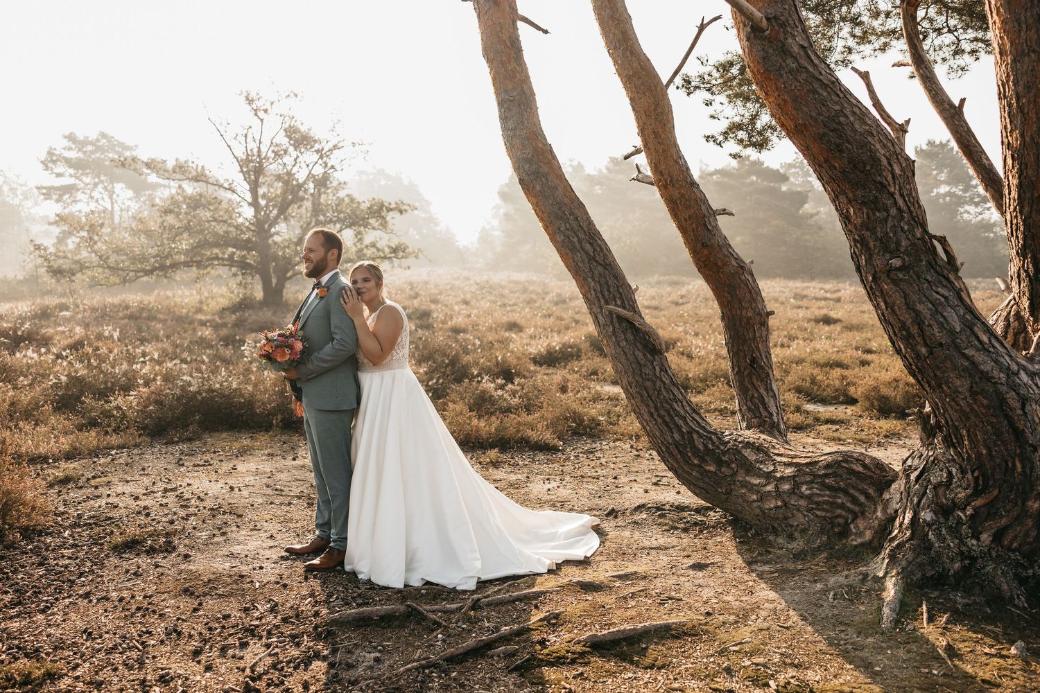Newlyweds pose together in a sunlit misty forest, bride in white gown and groom in grey suit with bouquet.