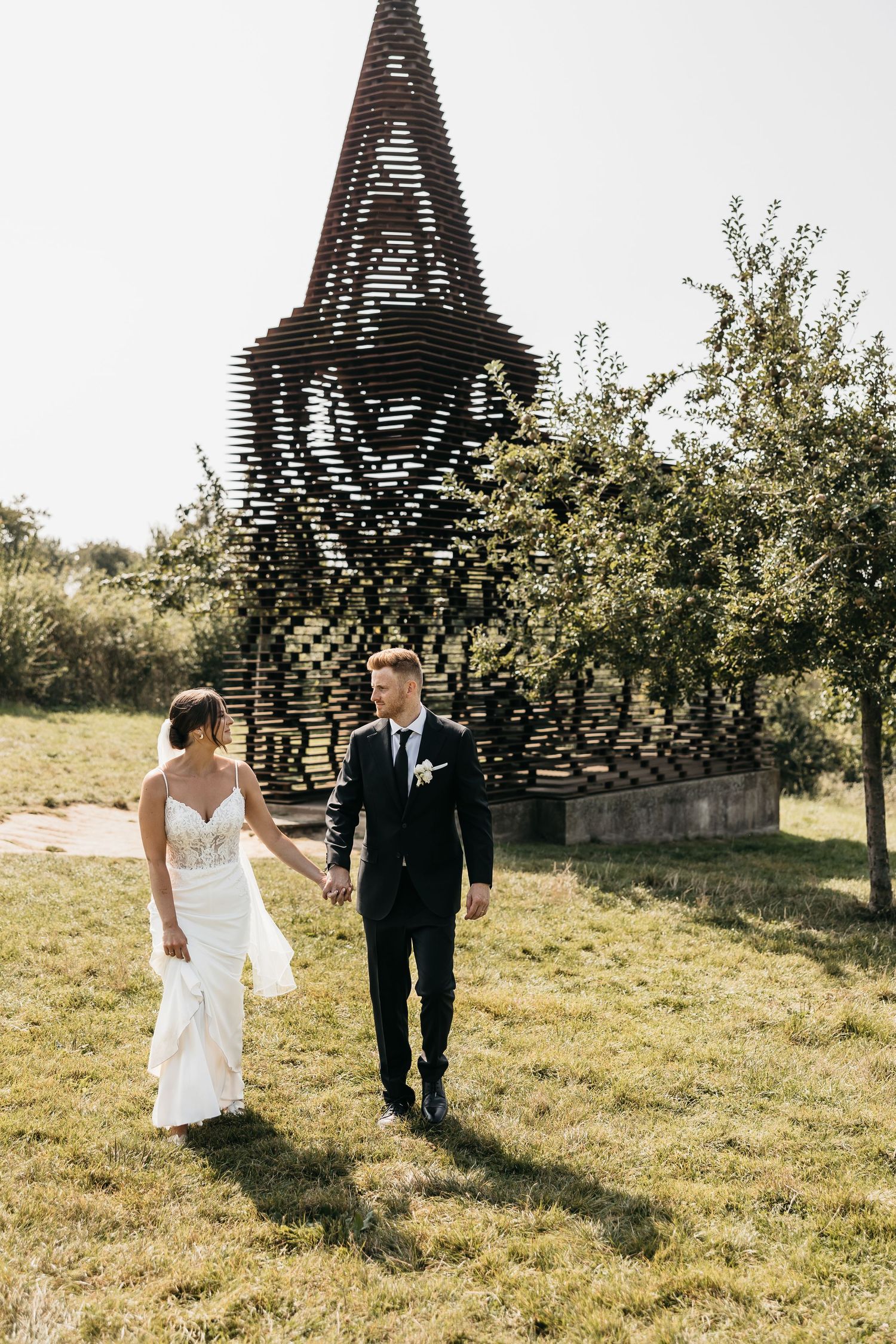 Bride and groom strolling hand in hand on sunny day near a decorative latticed church steeple.